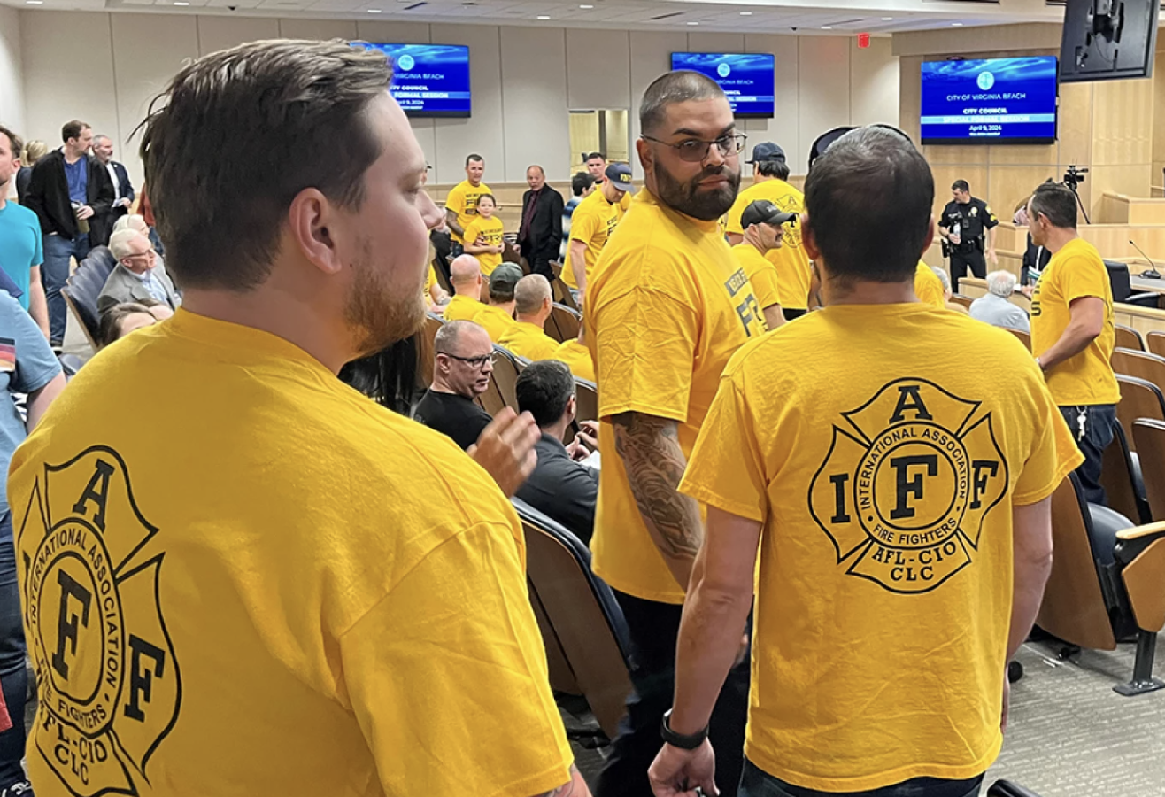 Group of people attending, many wearing yellow t-shirts with the fire fighters' logo on the back, in a large room with tiered seating and digital screens on the wall.