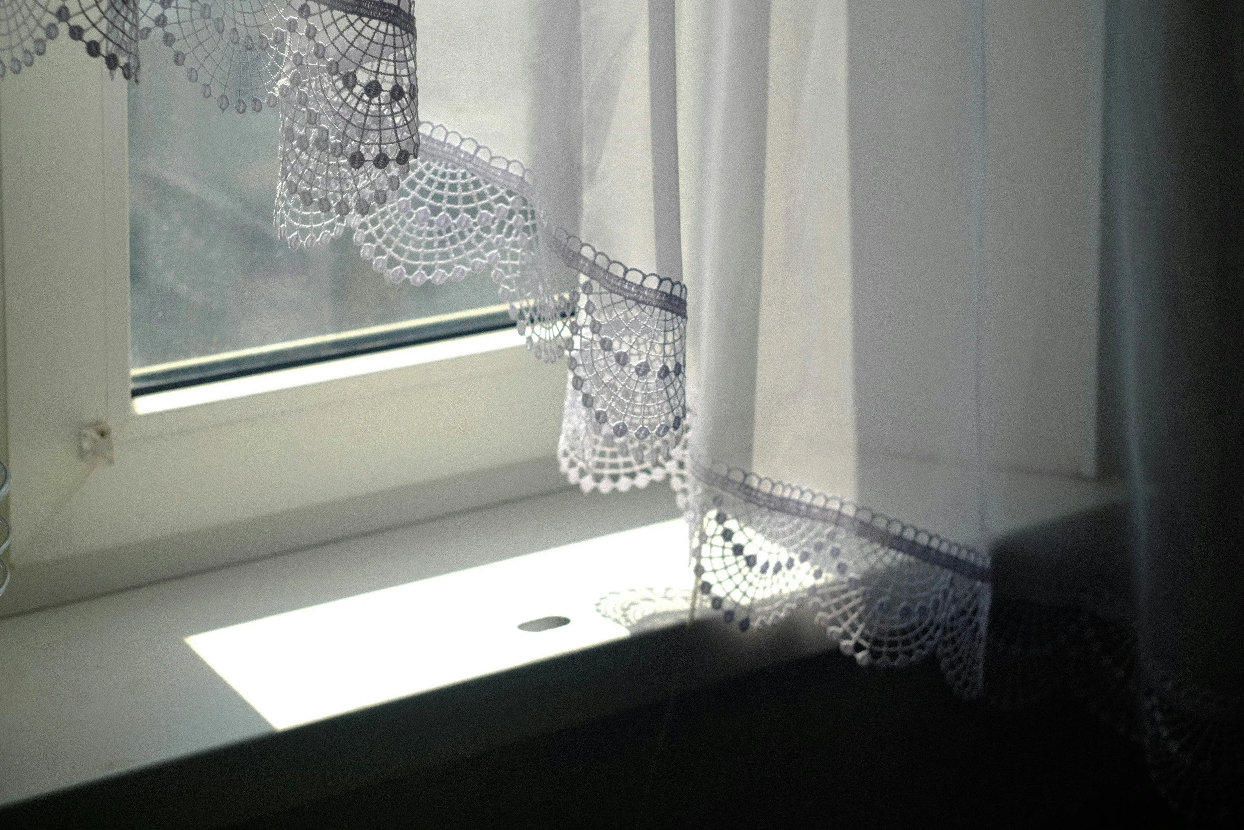 Sunlight coming through a window with a white lace curtain that has a scalloped edge and intricate crochet patterns.