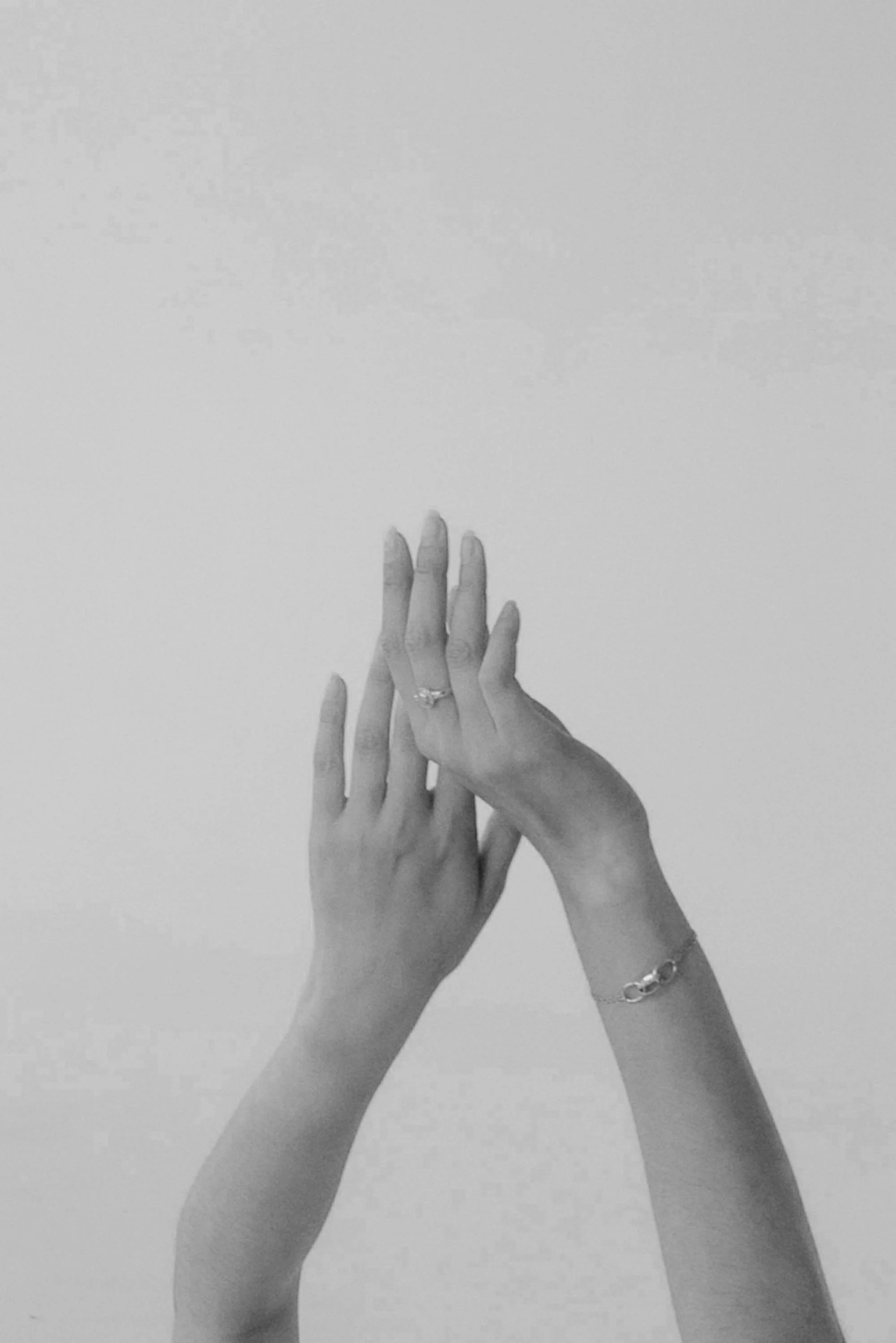 Two hands with rings and bracelets touching against a plain background, black and white photograph.
