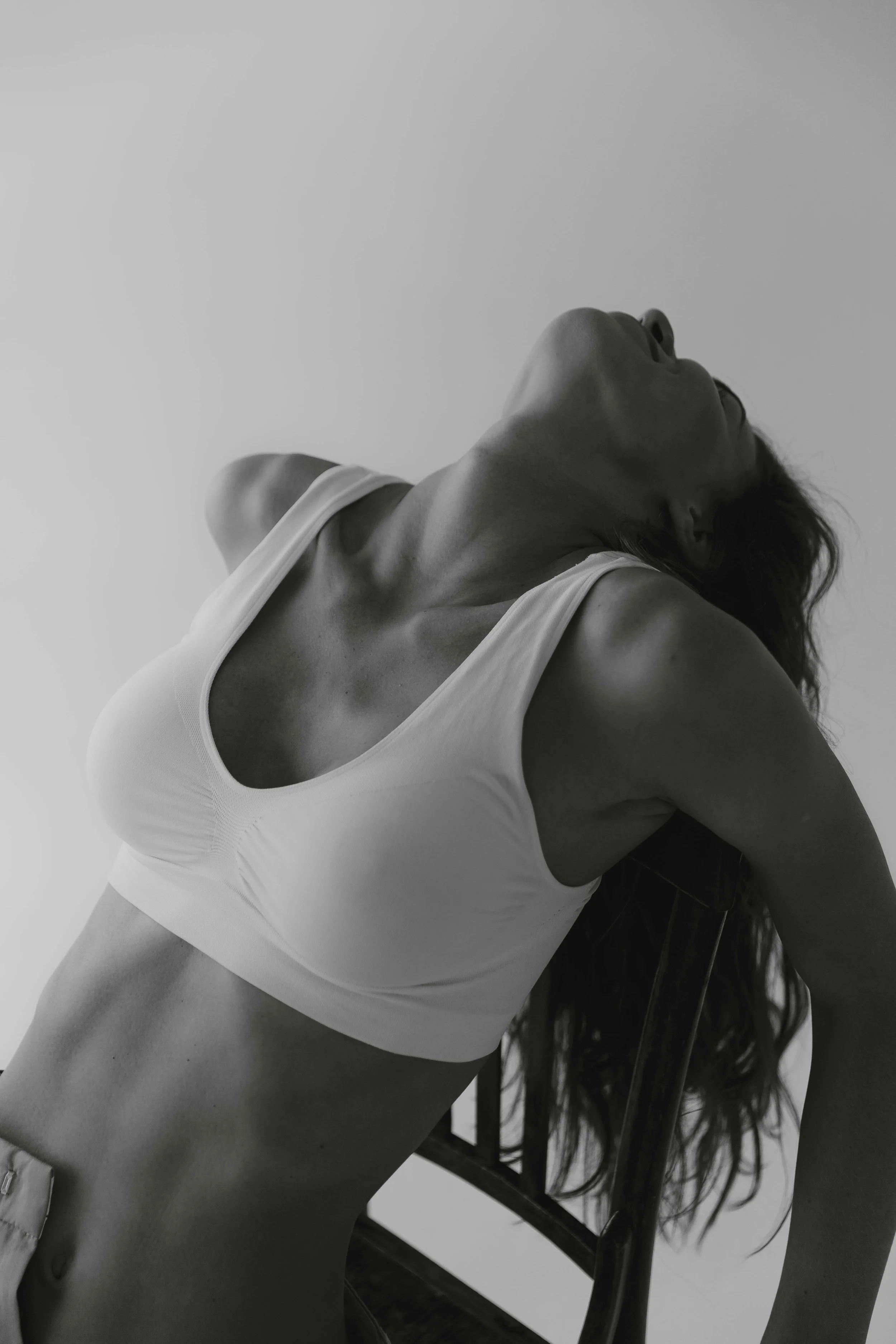 A woman in a white sports bra leaning back with her eyes closed, hair hanging down, and one arm resting behind her head, in black and white.