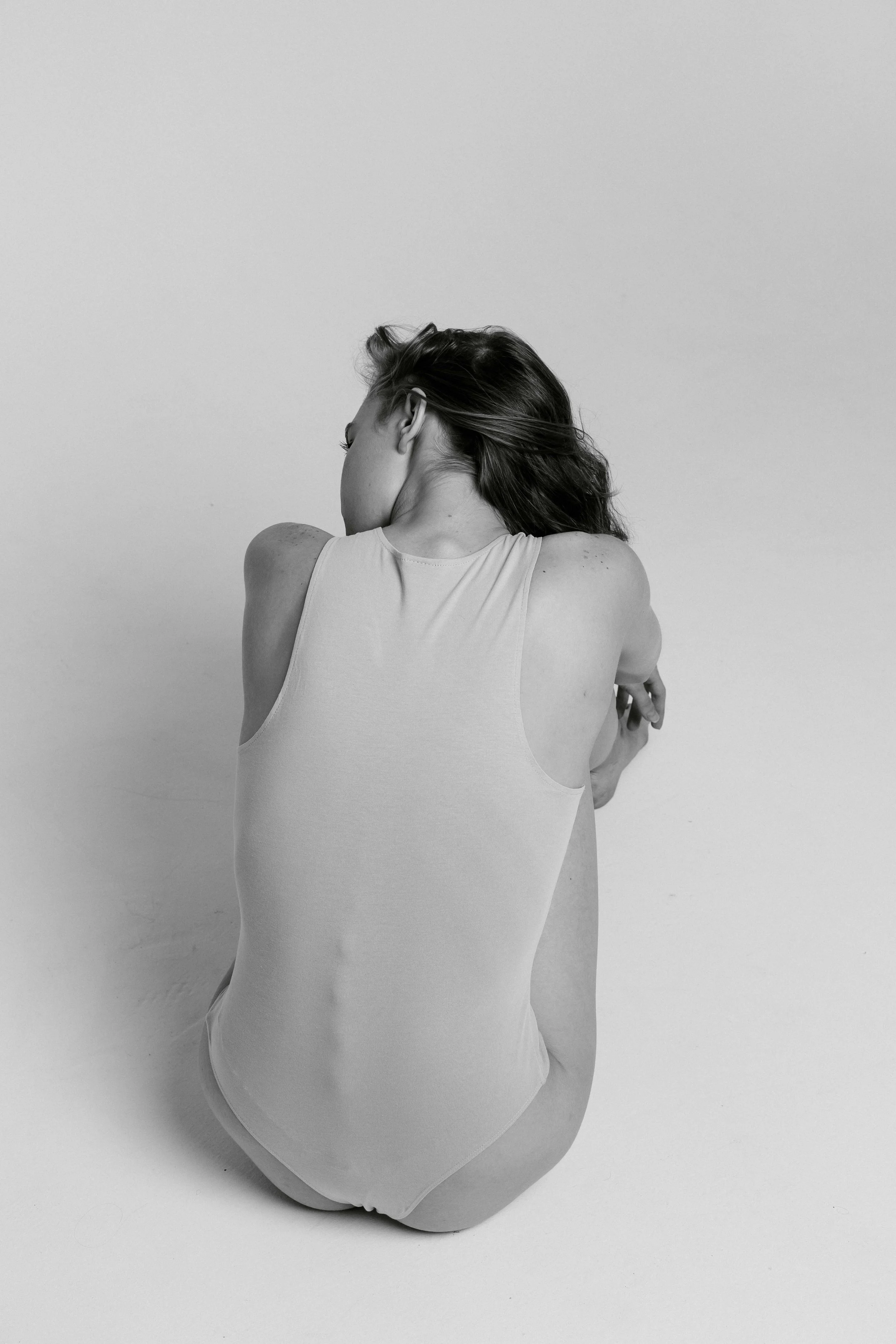 A woman with long hair wearing a tank top and underwear, sitting on the floor with her back to the camera, against a plain white background.