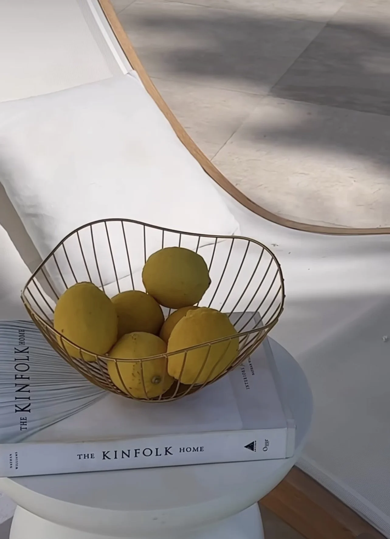 A wire bowl containing yellow lemons placed on top of two books titled 'The Kinfolk Home' on a white surface with a pillow and a mirror in the background.