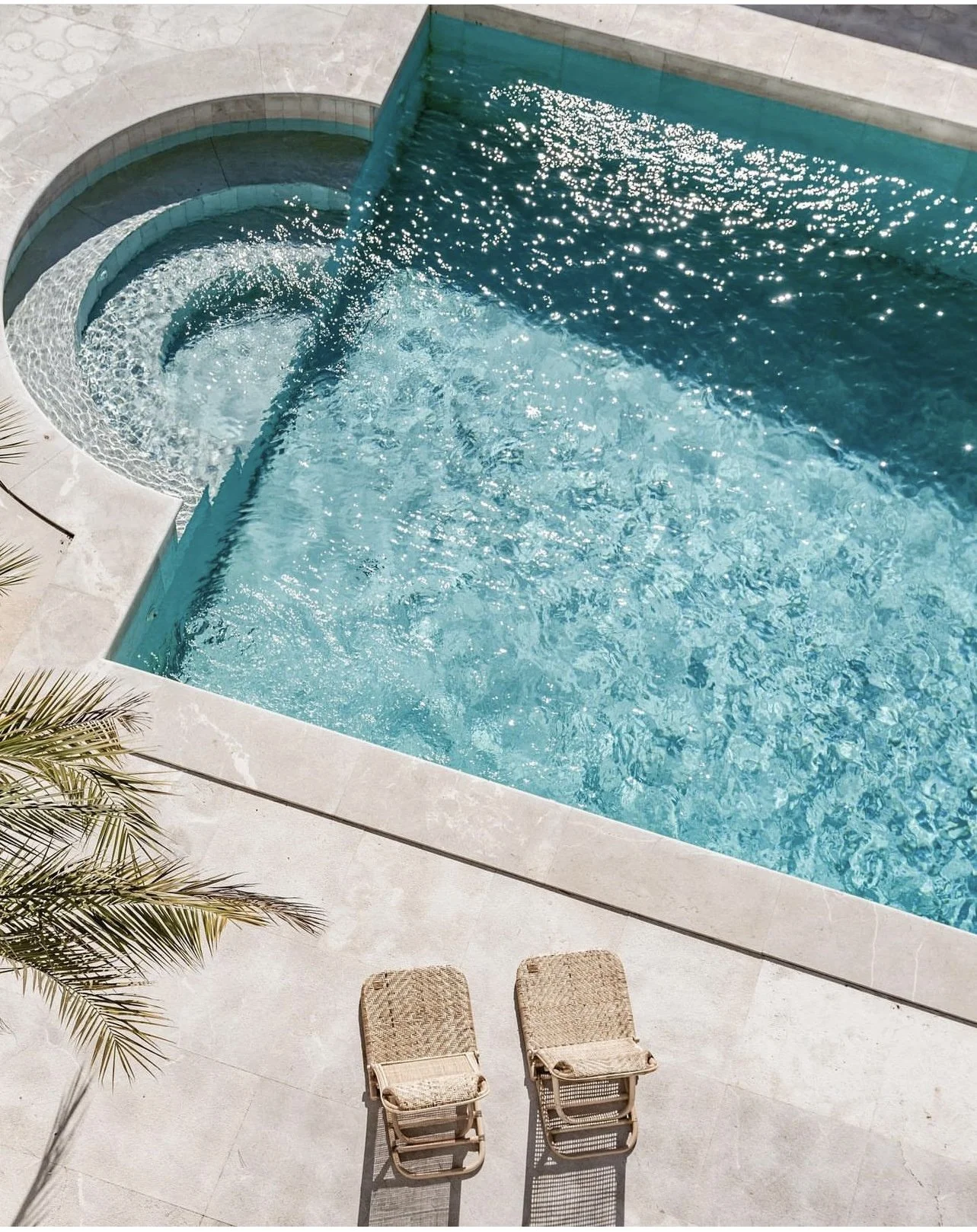 An aerial view of a backyard swimming pool with two lounge chairs on the beige stone deck, a small palm tree on the left, and a cascading water feature with water flowing into the pool.