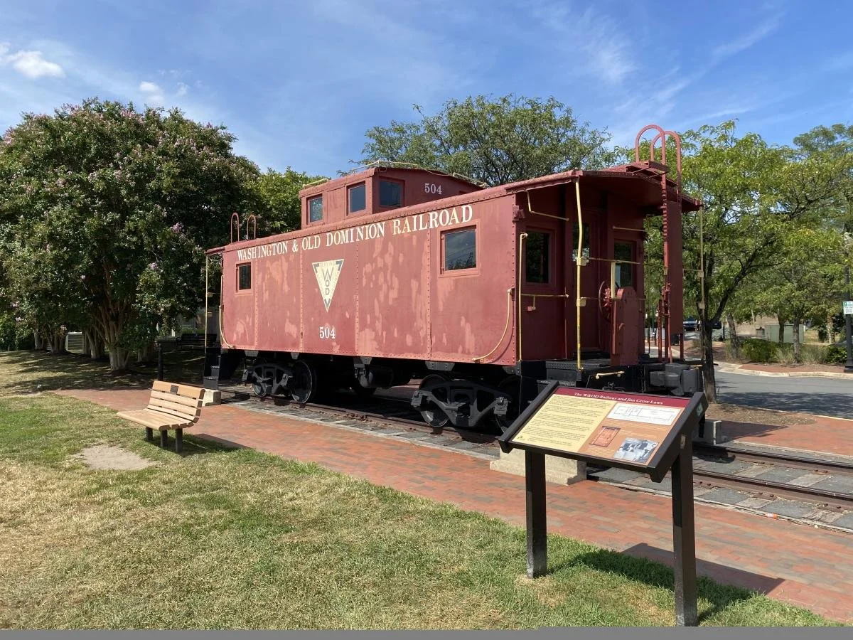 A vintage red train caboose with the words 'Washington & Old Dominion Railroad' written on the side, displayed outdoors on a small section of railroad track. There is an informational sign in front of it, a wooden bench nearby, and trees in the background.