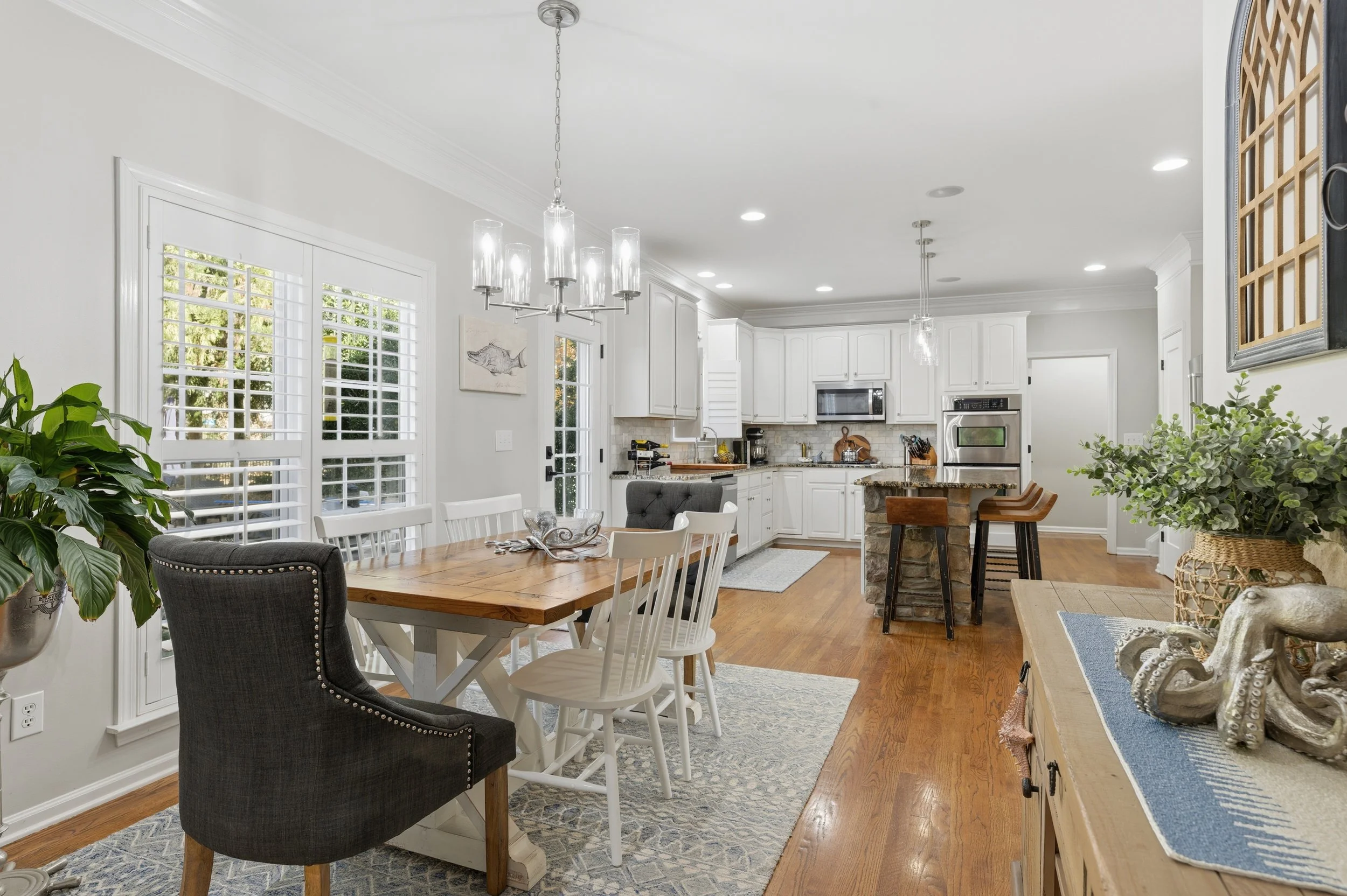 Bright, open-concept kitchen and dining area with white cabinetry, hardwood floors, and decor that includes plants, a fish artwork, and nautical-themed ornaments.