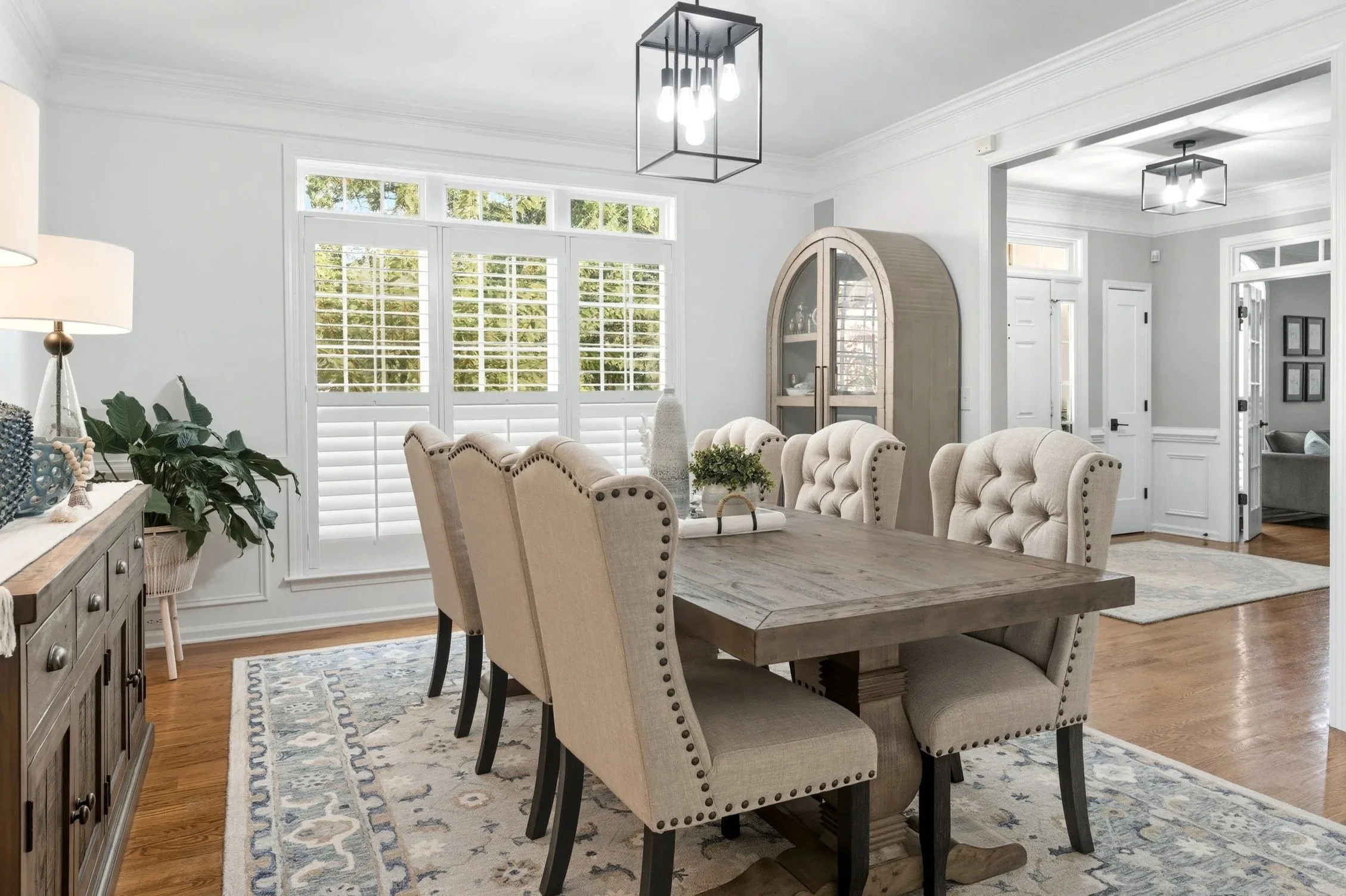 Bright dining room with large window, wooden table, beige upholstered chairs, and a patterned rug.