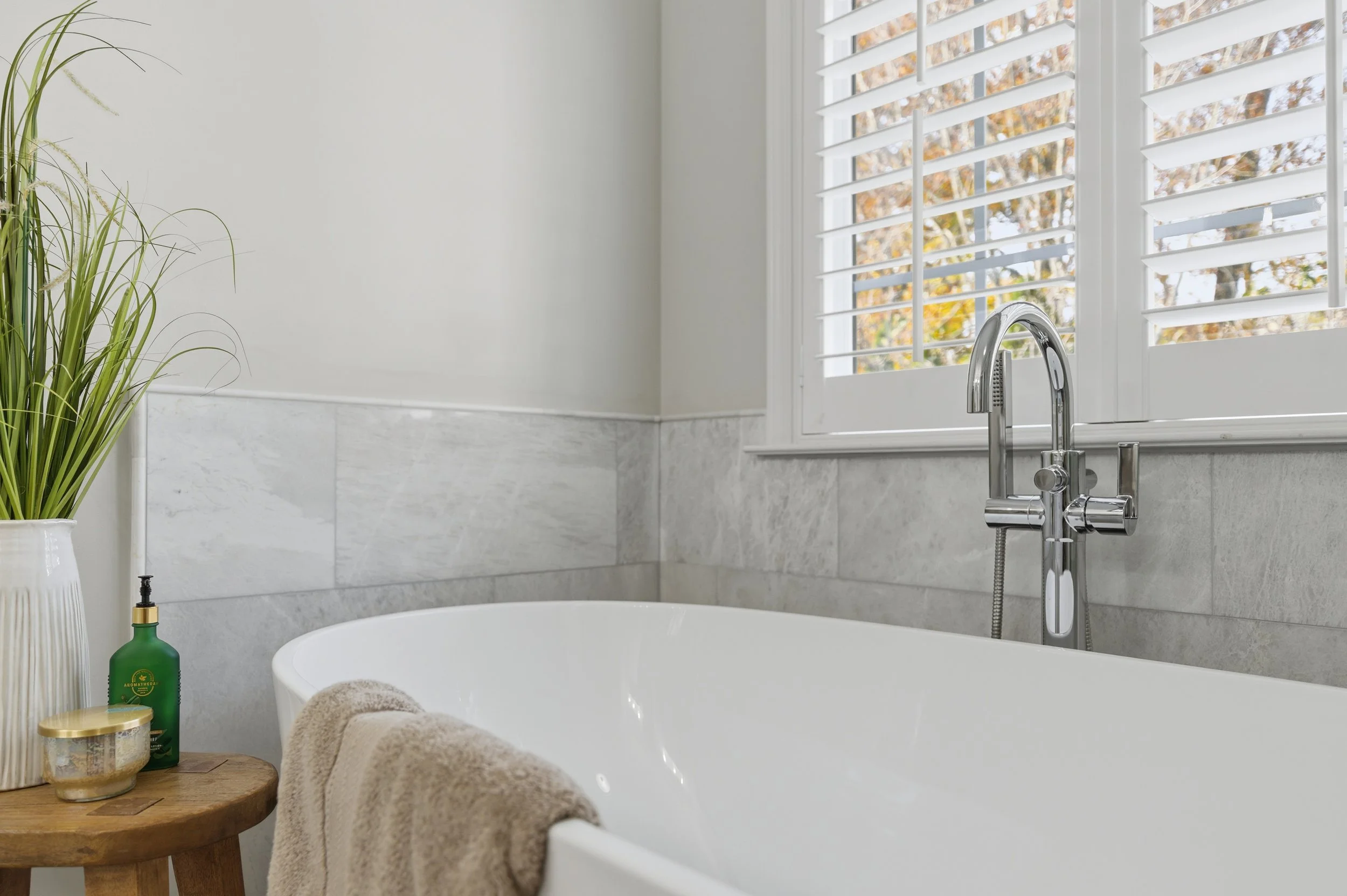 Bathroom with white freestanding bathtub, window with white plantation shutters, green soap bottle, gold-accented candle, and a potted plant on a wooden stool.