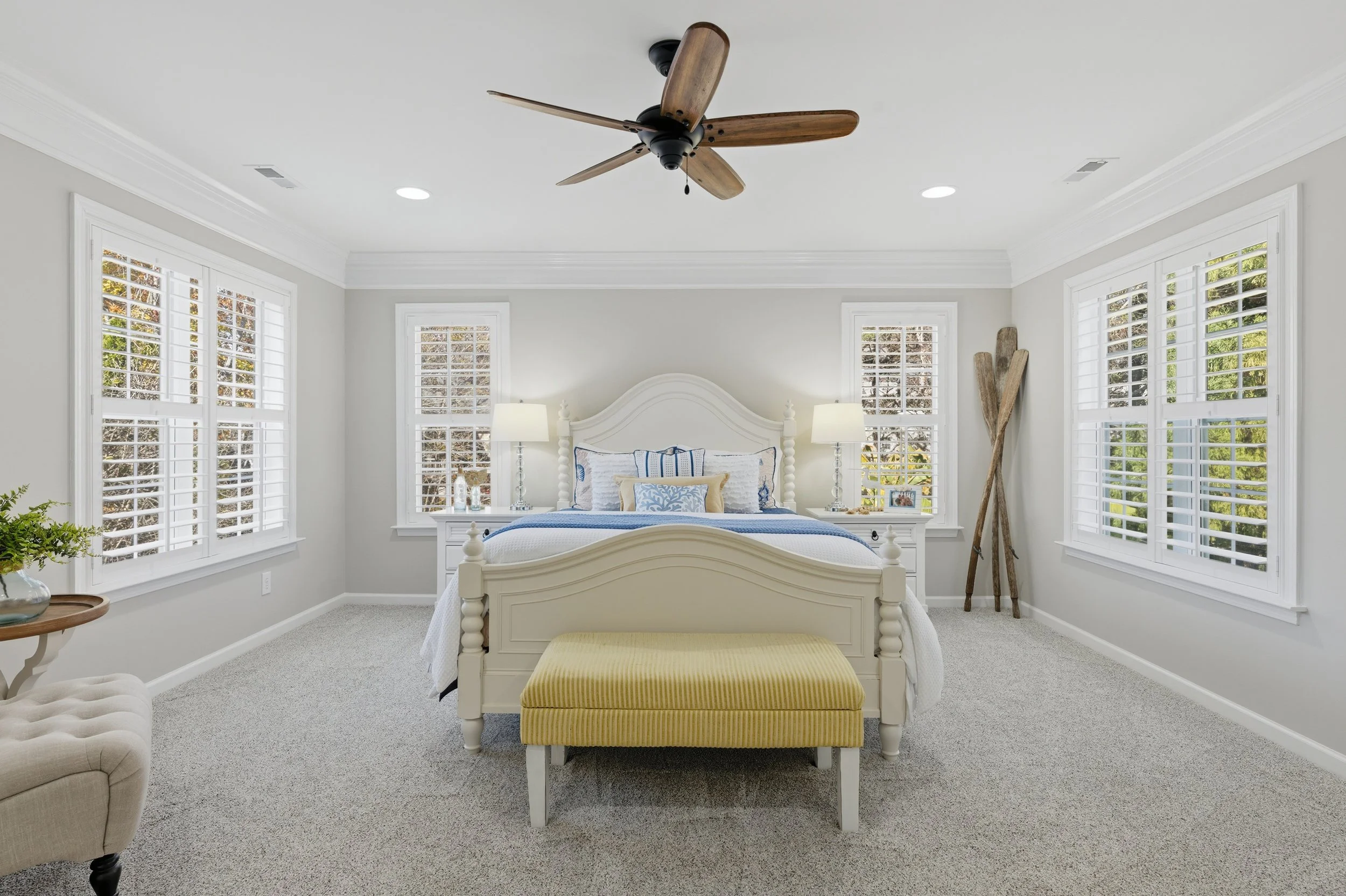 Bright, spacious bedroom with a white bed, yellow bench, and large windows with white shutters.