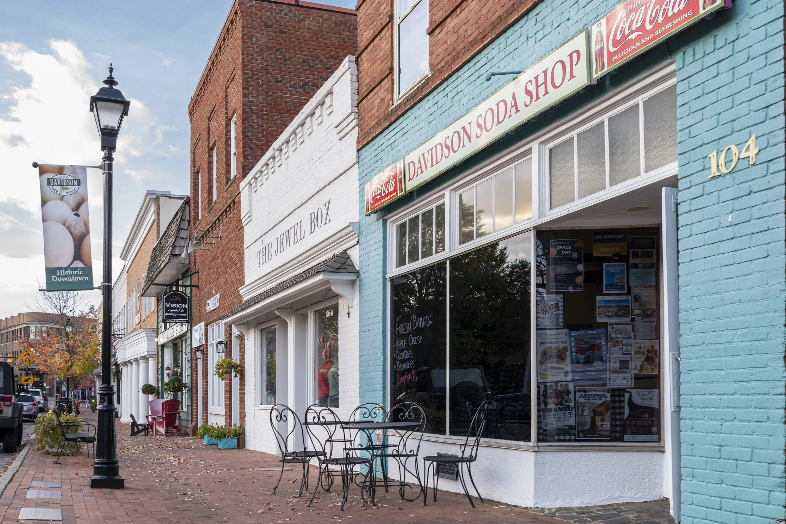 Street view of a row of storefronts with outdoor seating, a lamppost, and a banner. The storefronts include a soda shop with a Coca-Cola sign and a jewelry box shop. The sky is partly cloudy.