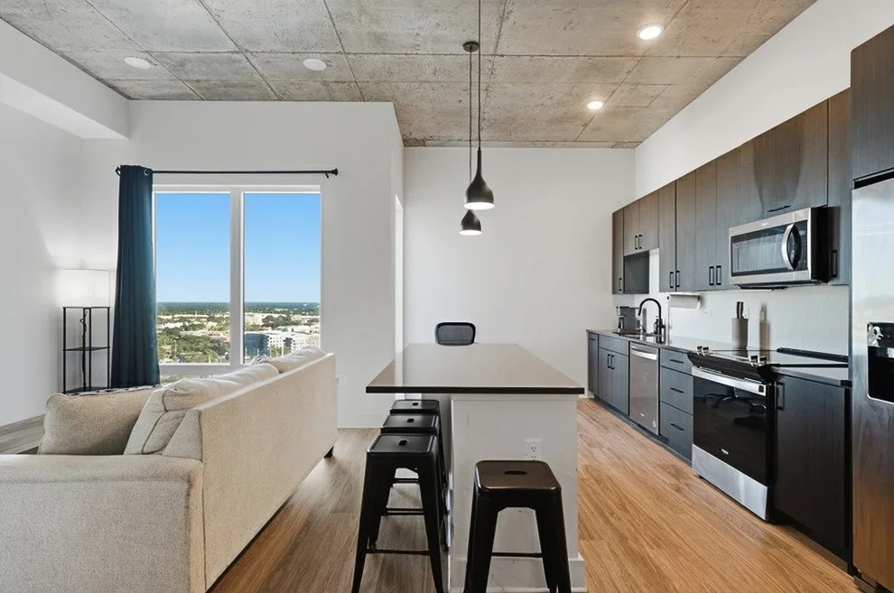 Modern kitchen and living room with large window view of city, beige sofa, black bar stools, dark wood cabinets, stainless steel appliances, and minimal decor.