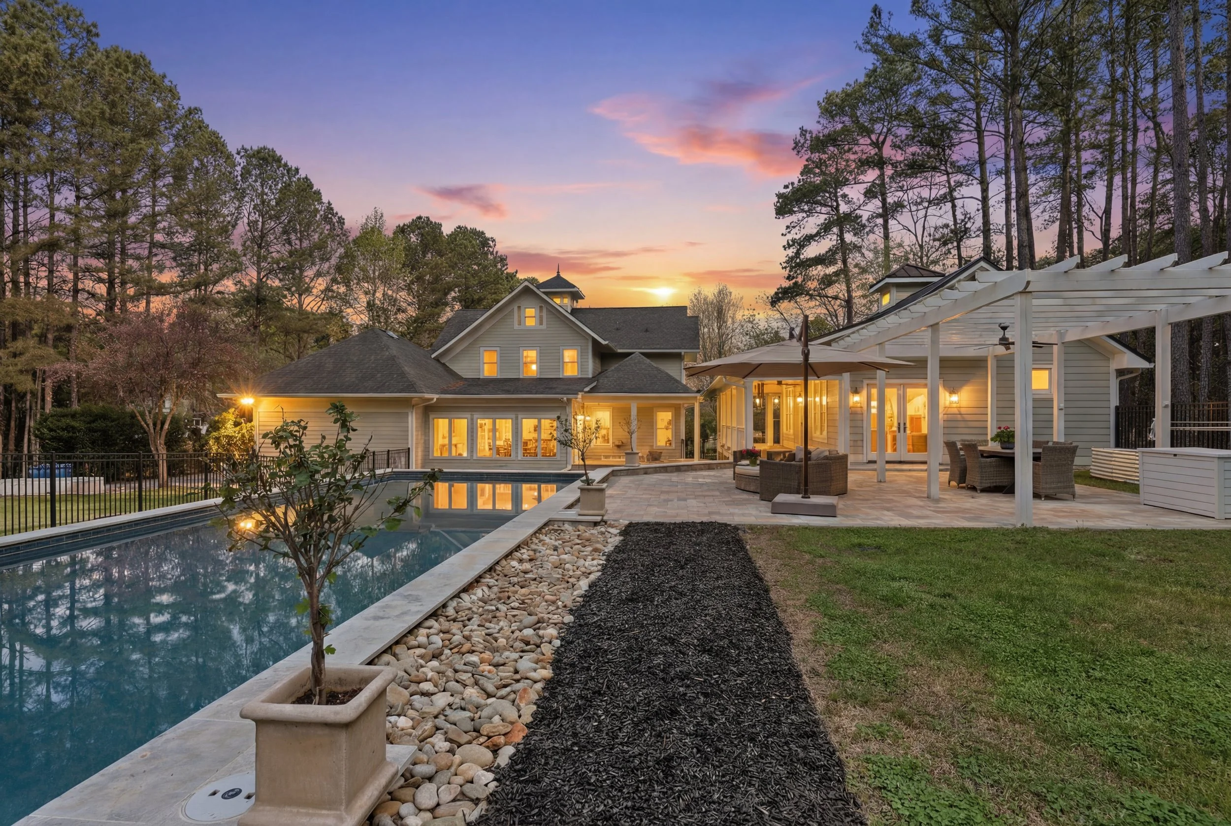 A large brick house with black shutters, a white front porch with stairs, and a garage, surrounded by fallen autumn leaves and trees with orange and green foliage at sunset.