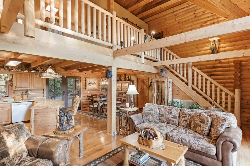 Interior of a cozy log cabin living room with wood-paneled walls and ceiling, featuring a patterned upholstered sofa, stone fireplace, and a loft level with white wooden railing.