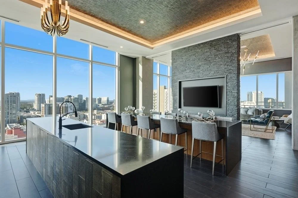 Modern high-rise apartment with an open kitchen and living area featuring large windows with city views, a black kitchen island, a wall-mounted flat-screen TV, and contemporary furniture.
