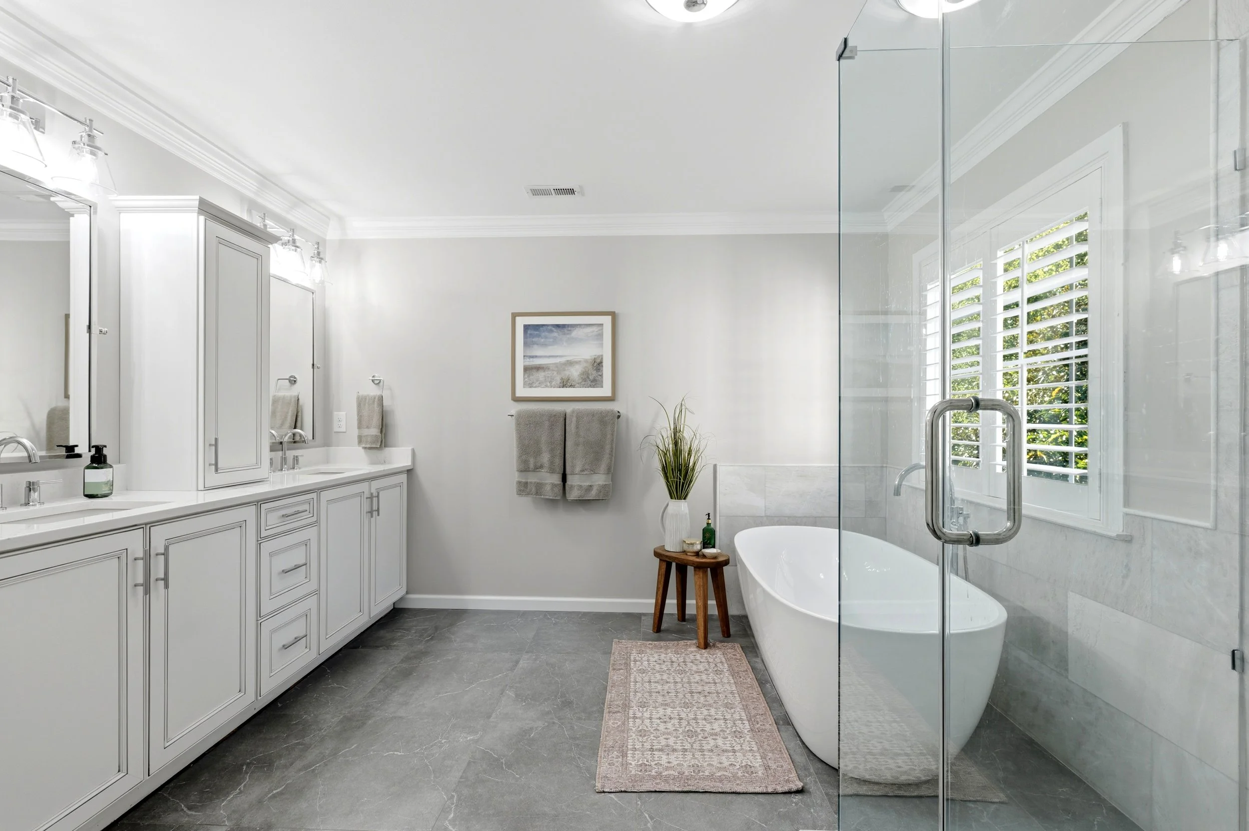 Modern bathroom with white vanity, large mirror, gray tiles, bathtub, and window with shutters