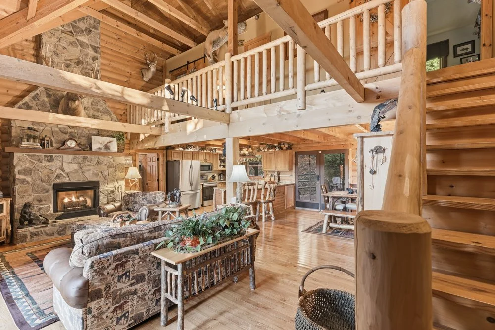 Cozy log cabin living room with stone fireplace, wooden furniture, and loft area with animal mounts, wood staircase, and large windows.