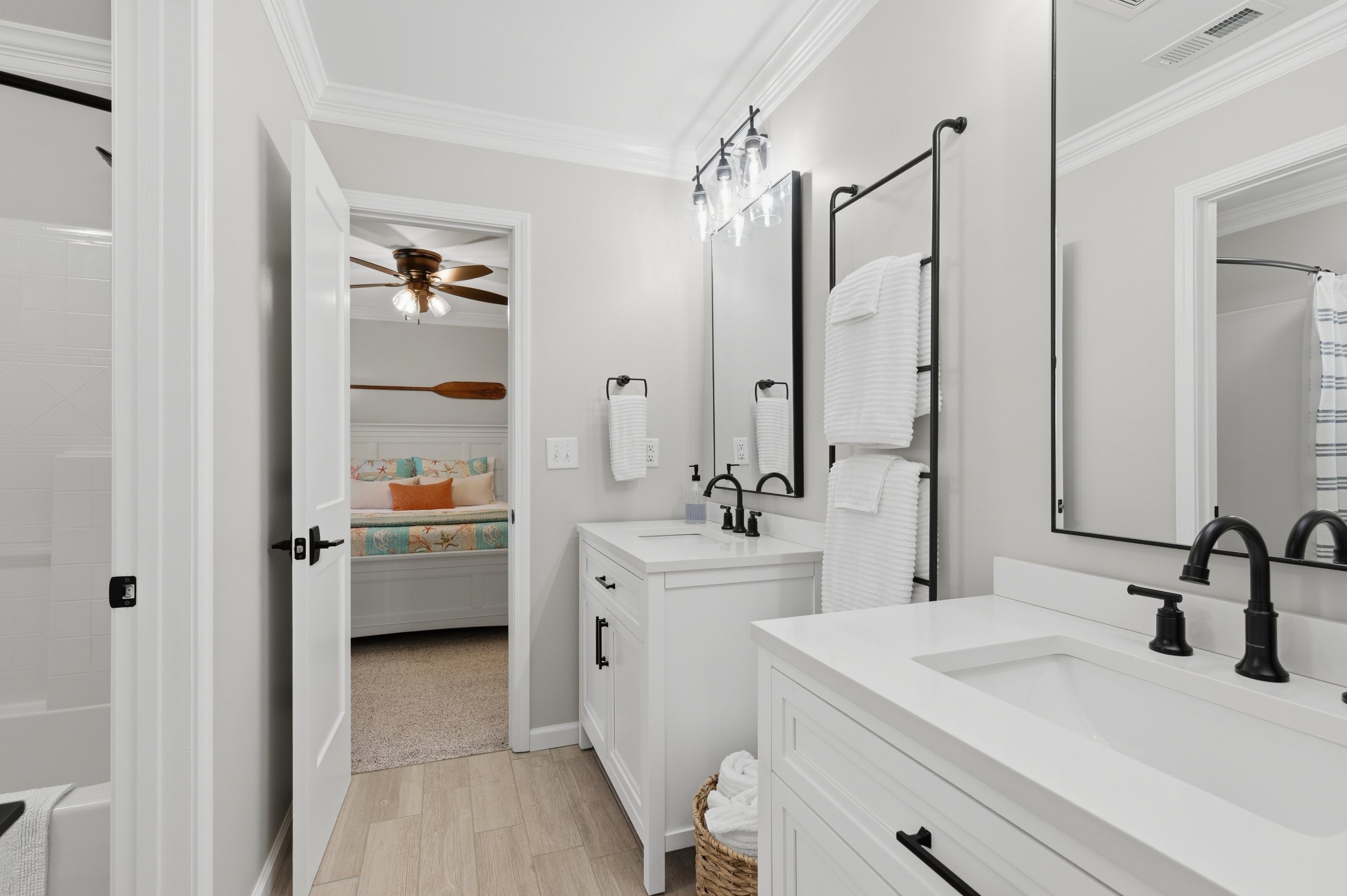 Modern bathroom with white vanity, black fixtures, large mirrors, and towel racks, overlooking a bedroom with a bed and a ceiling fan.