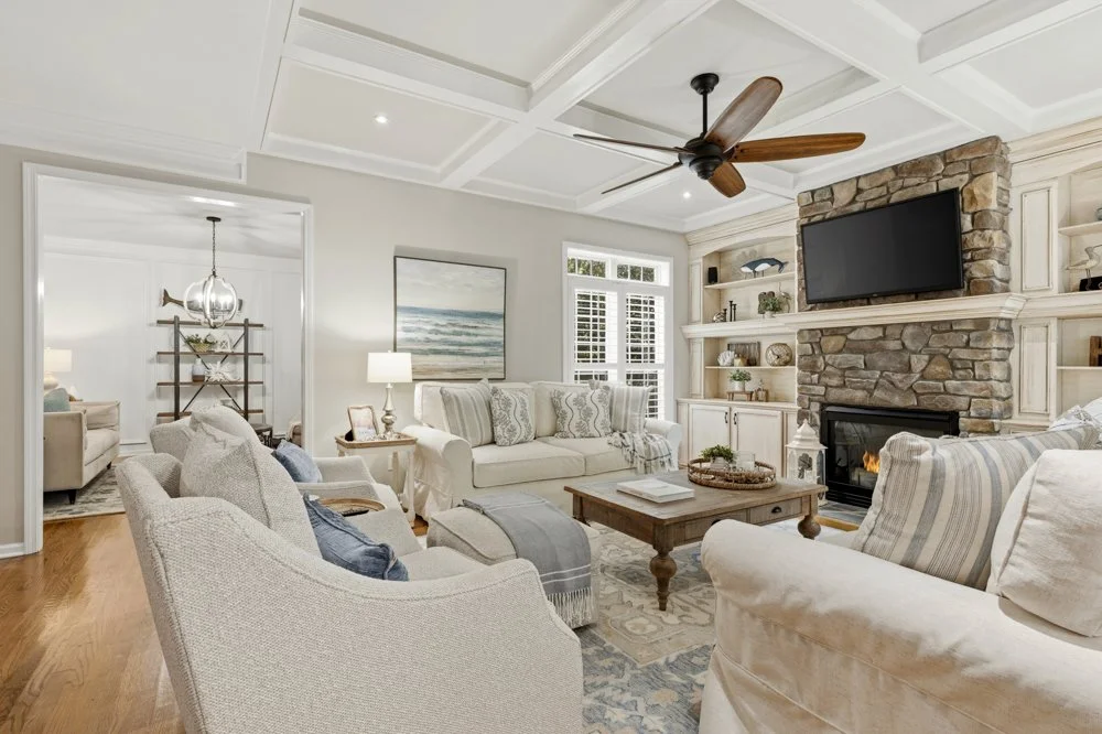 Bright living room with white walls, a stone fireplace, a flat-screen TV, and white and beige sofas, with a wooden coffee table and ceiling fan.