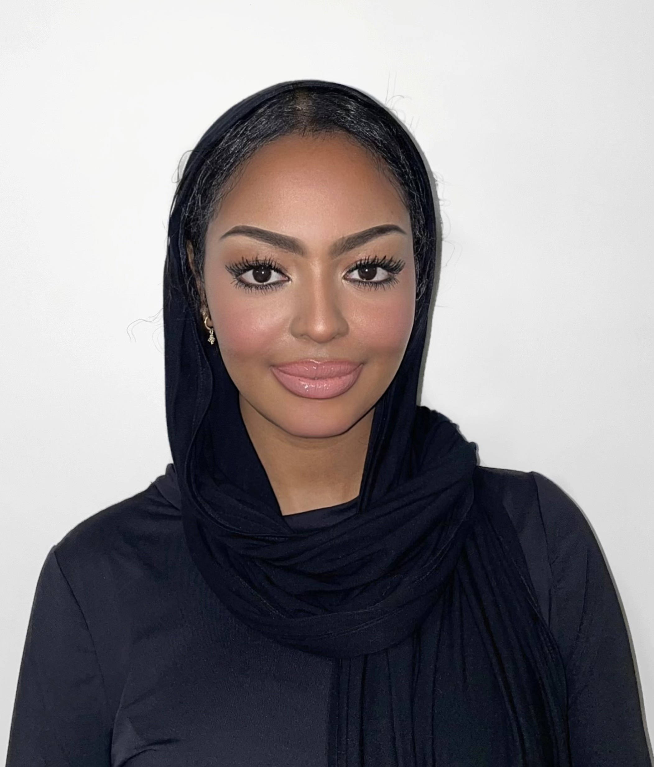 A woman with medium skin tone, dark hair, wearing a black headscarf, black top, and gold earrings, smiling at the camera against a plain white background.