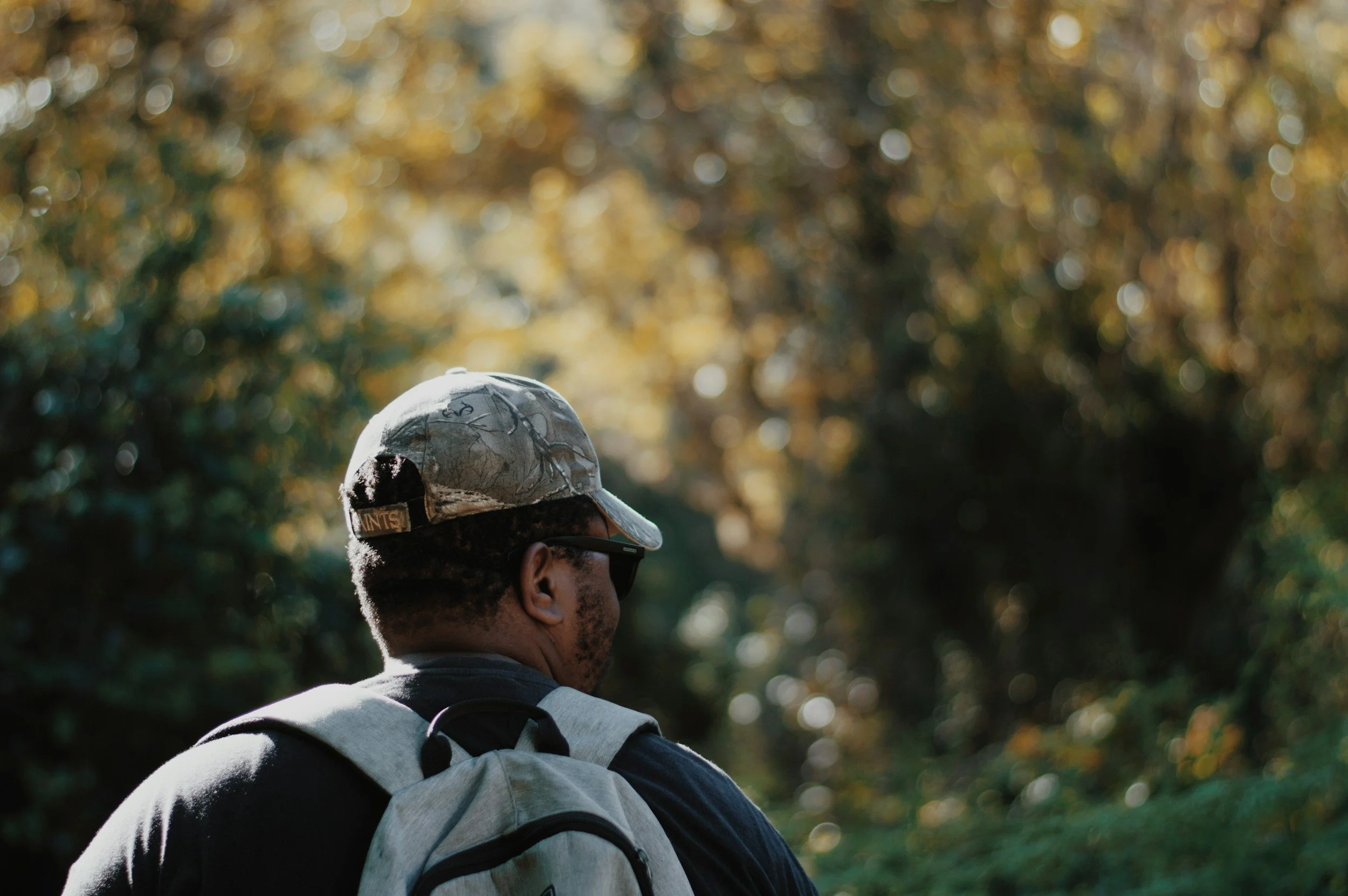 Man with a backpack and camouflage hat walking in a wooded area during daylight.