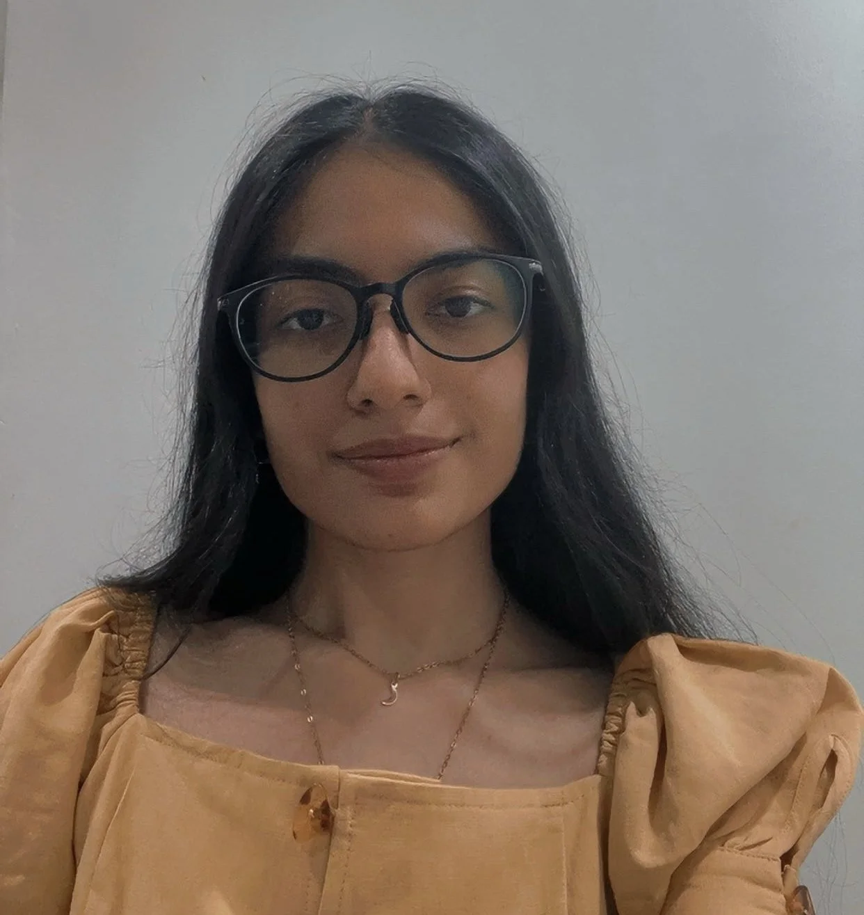 A young woman wearing glasses and a yellow top, smiling at the camera against a plain background. She has dark hair and is wearing two layered necklaces, one with a crescent moon pendant.