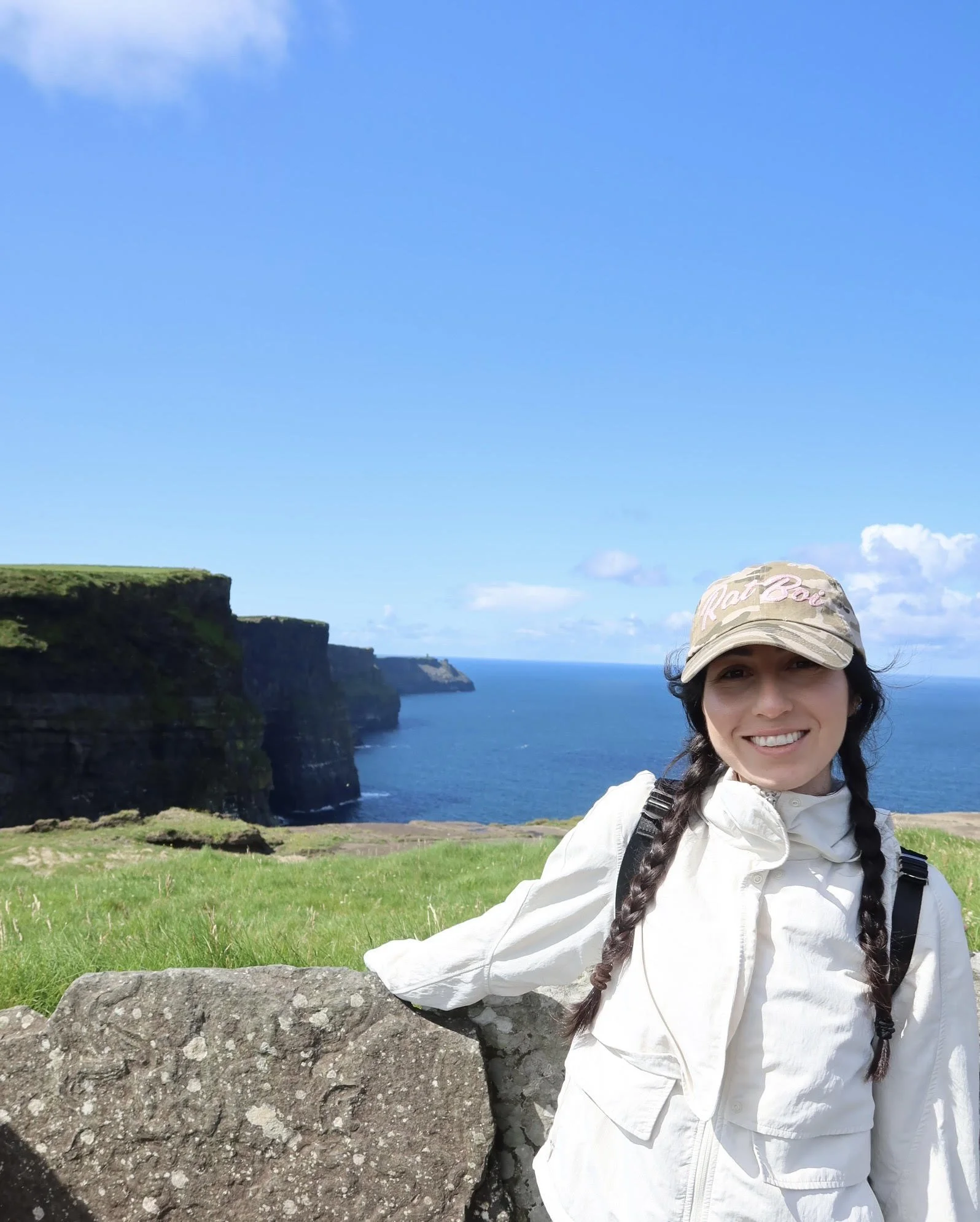 A woman smiling outdoors with green cliffs and the ocean in the background.