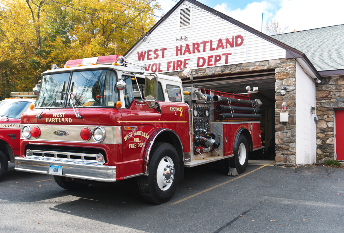 A vintage red West Hartland Volunteer Fire Department fire truck parked outside a fire station with trees in the background.