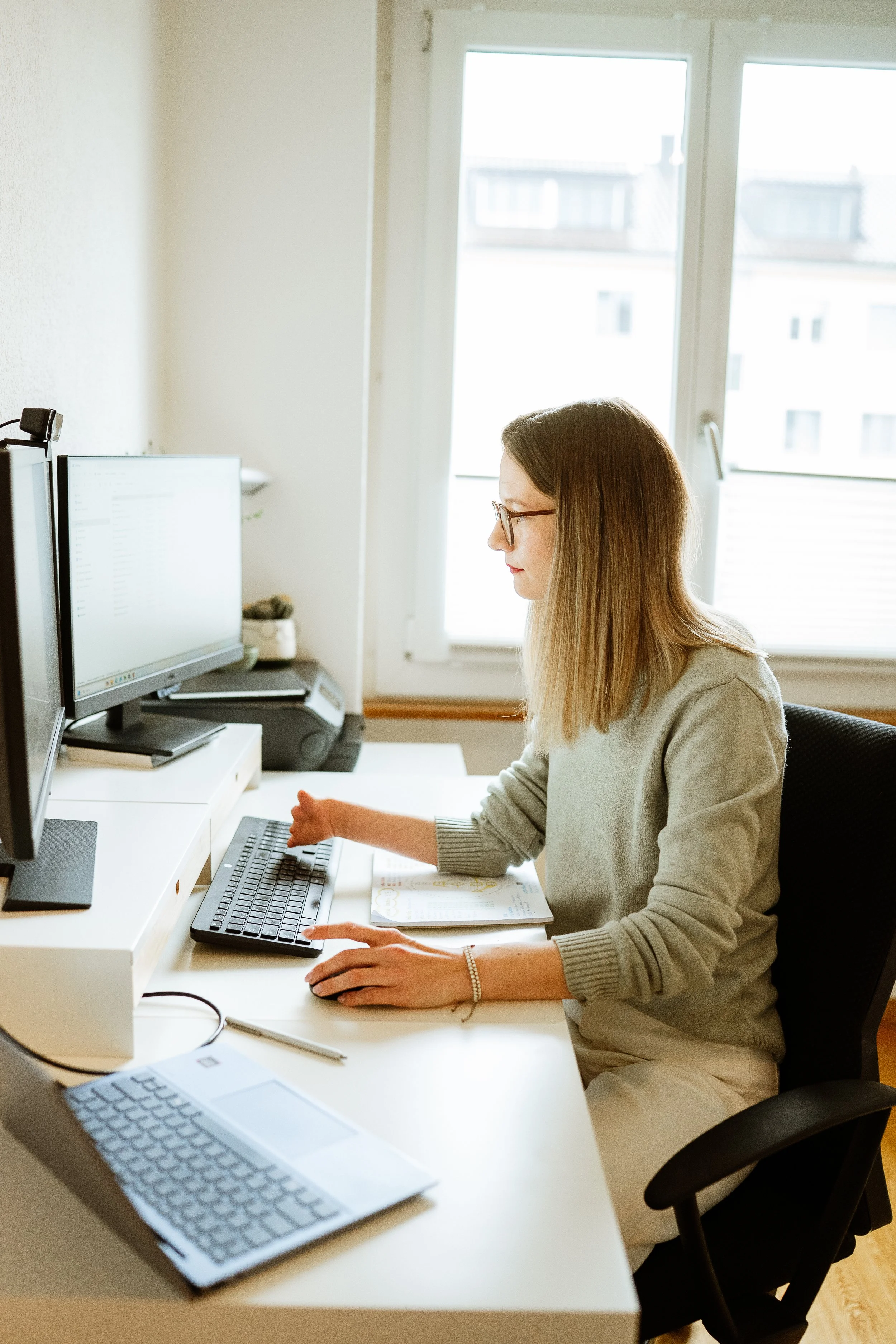Frau arbeitet an einem Schreibtisch mit zwei Monitoren, einer Tastatur, einem Laptop und Stift, in einem Raum mit großen Fenstern.