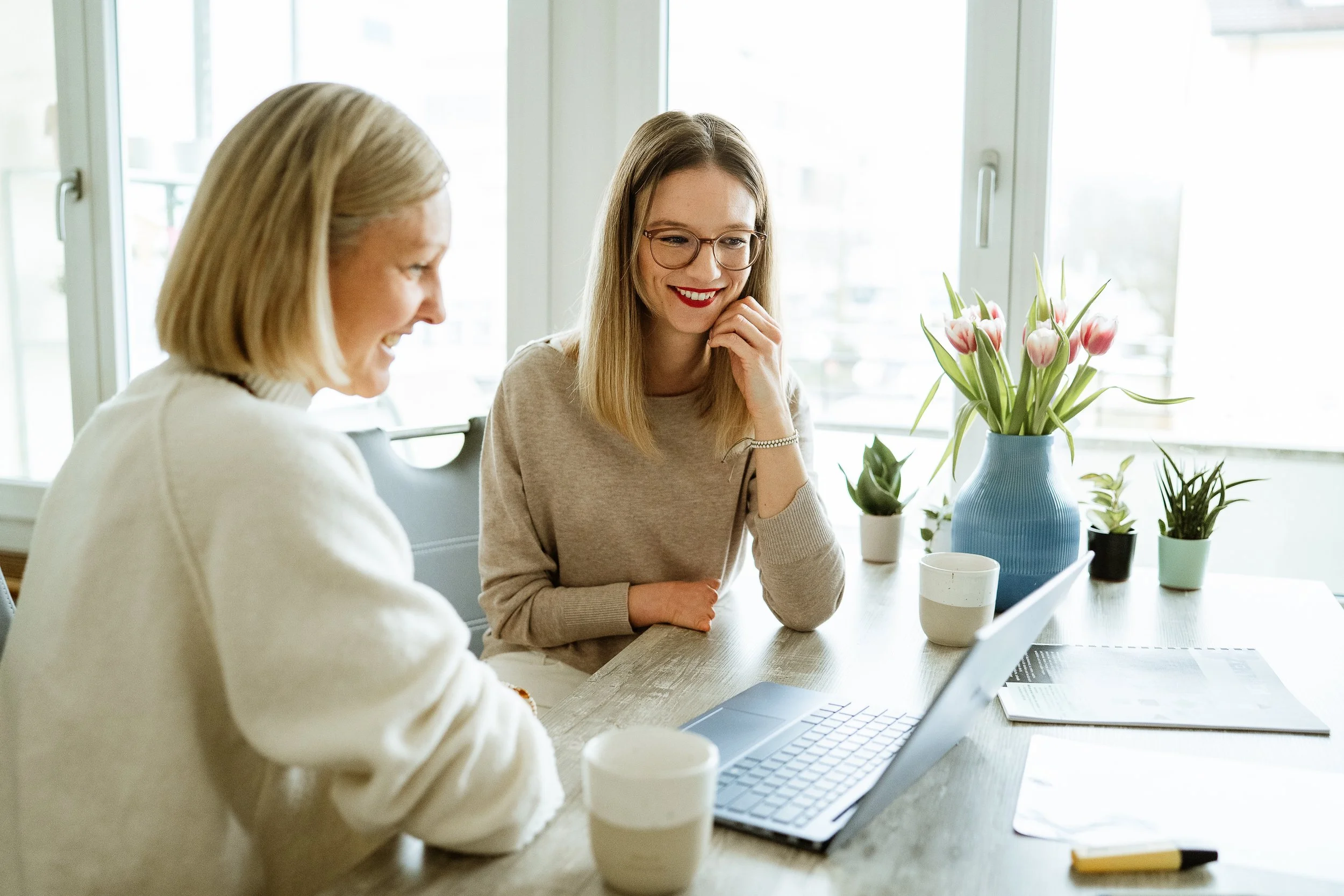 Zwei Frauen sitzen an einem Tisch mit einem Laptop und trinken Kaffee, während sie sich unterhalten. Im Hintergrund sind Fenster mit Blick auf eine Stadt und mehrere Topfpflanzen, darunter Tulpen in einer blauen Vase.