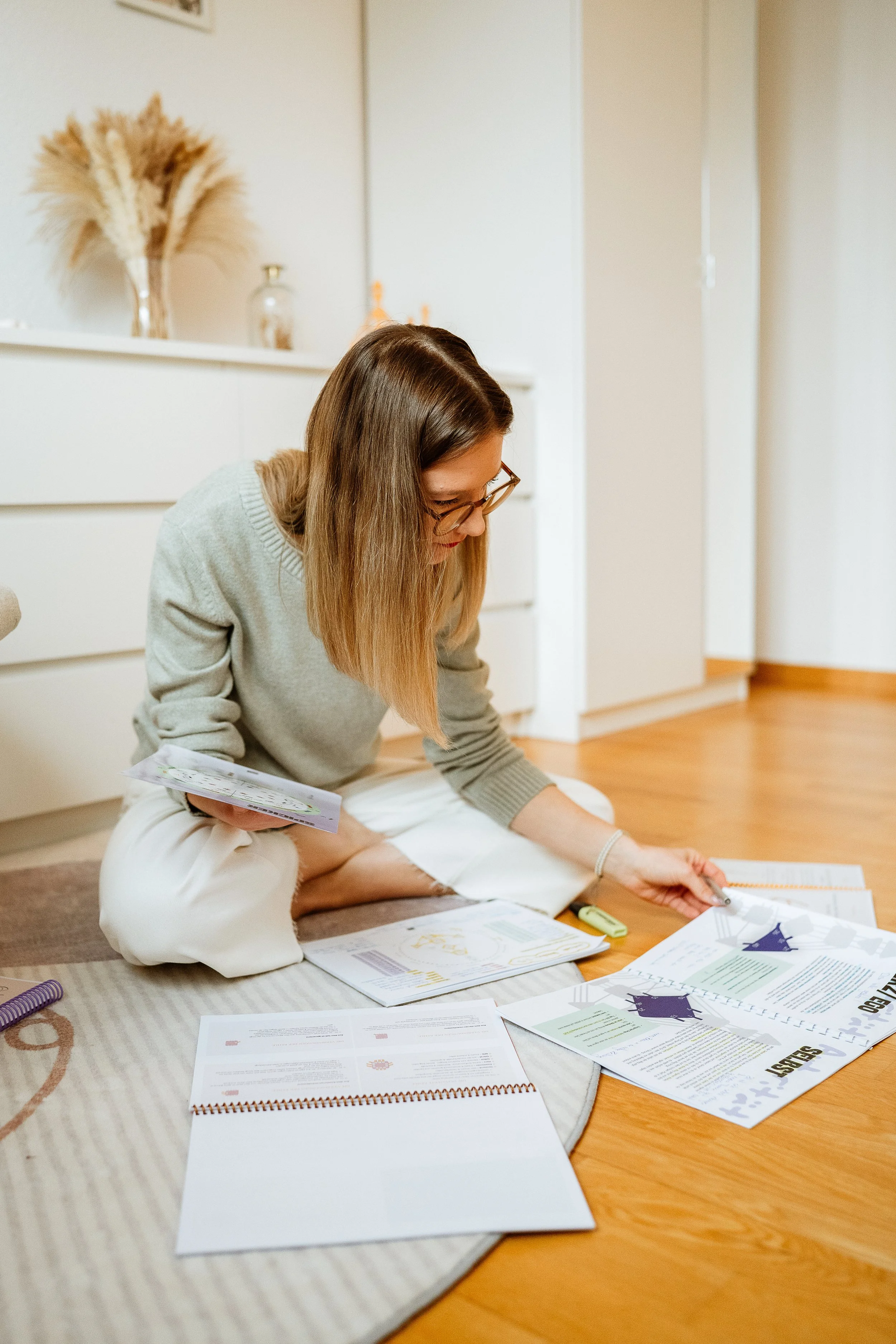 Eine Frau sitzt auf dem Boden und arbeitet mit Papieren und Notizen, während sie in einem Raum mit Holzboden und weißen Möbeln sitzt.