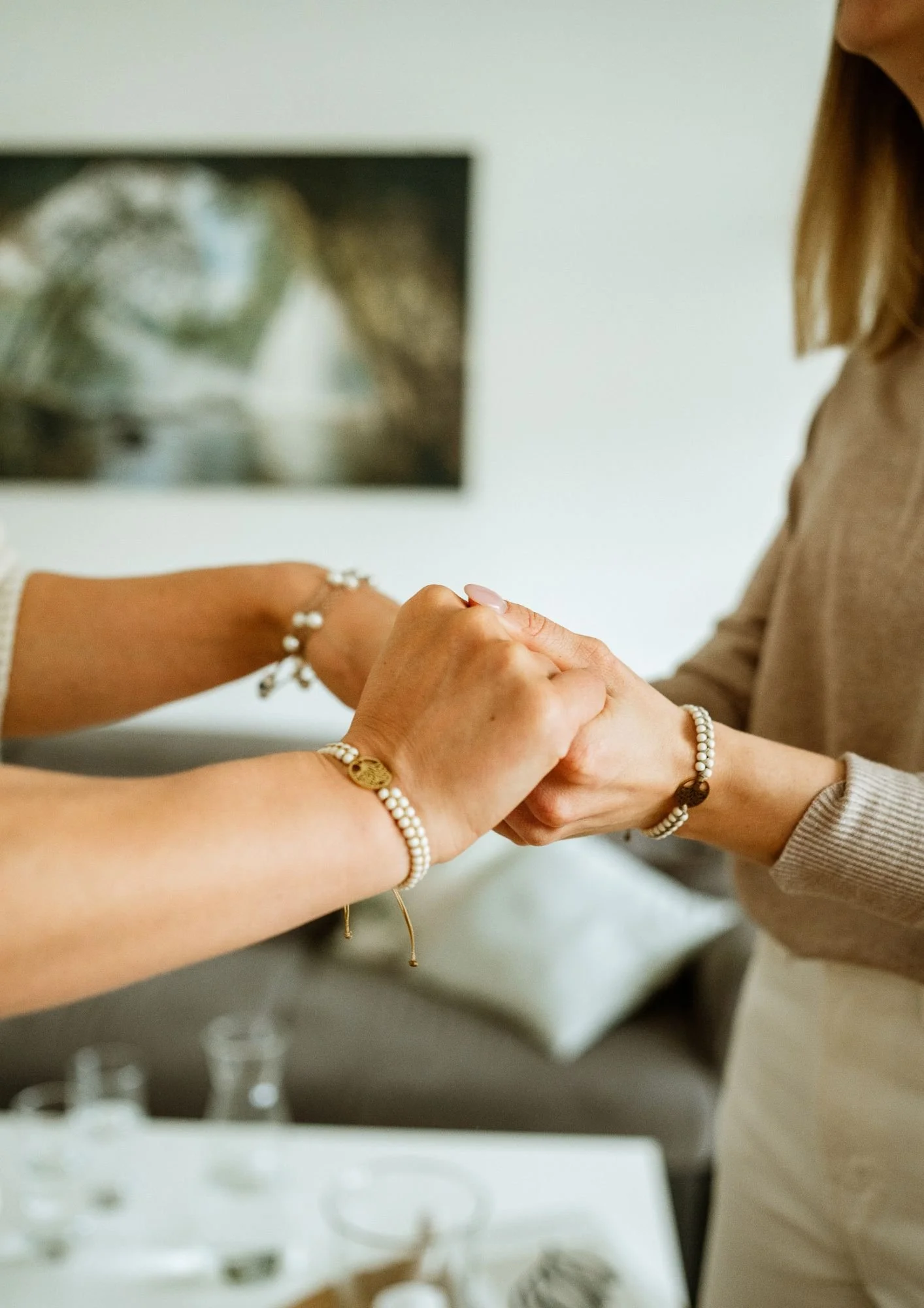 Zwei Frauen halten sich an den Händen in einem freundlichen Austausch, getragen von Schmuck und mit einem Bild an der Wand im Hintergrund.