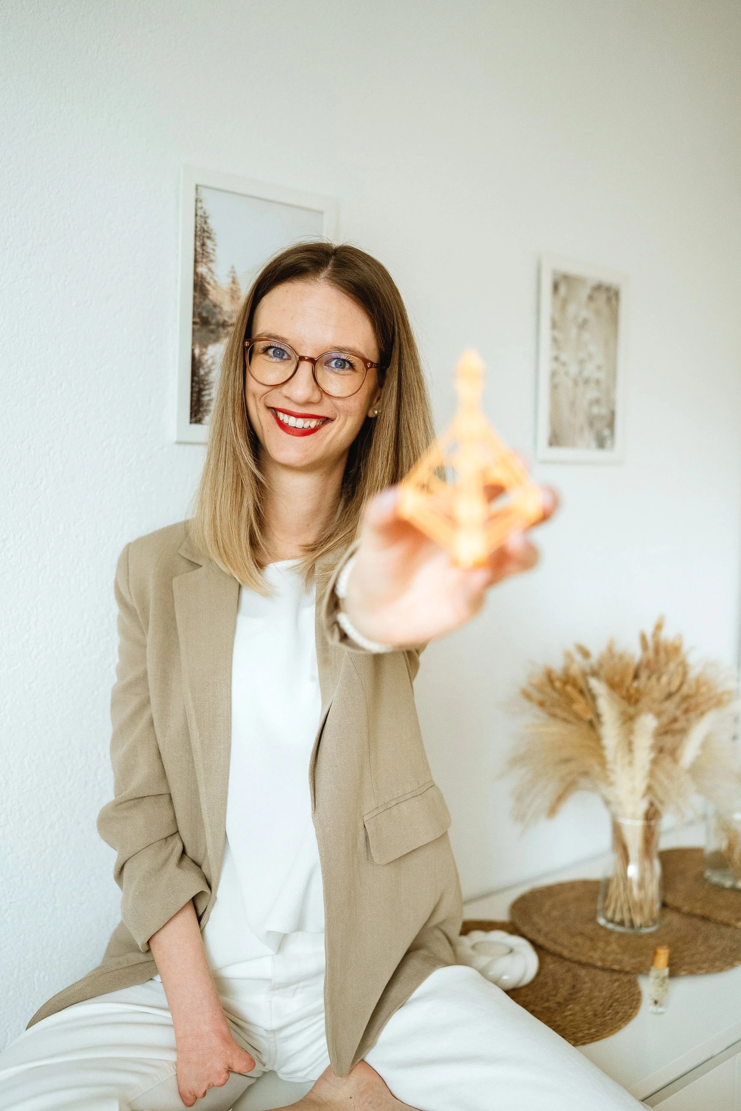 Lächelnde blonde Frau mit Brille hält eine kleine goldene Figur in die Kamera. Sie sitzt in einem hellen Raum mit Bildern an der Wand und einem Tisch mit Deko und Trockenblumen.