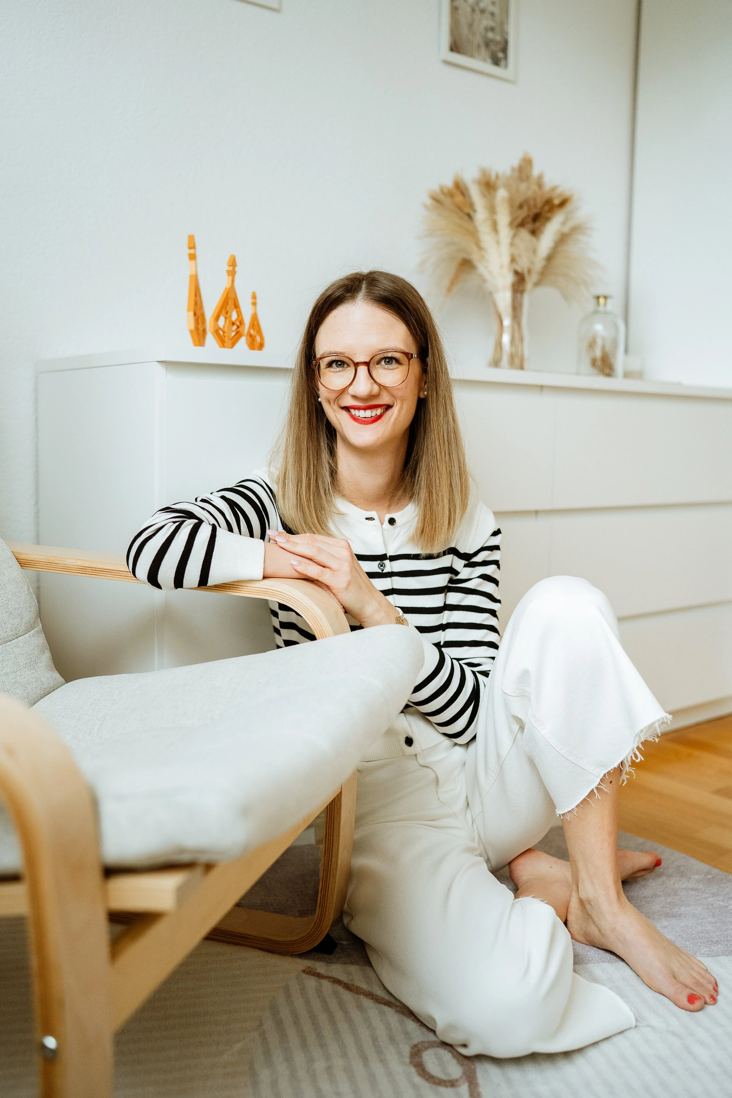 Eine lächelnde Frau mit Brille und langem Haar sitzt bequem auf dem Boden in einem modernen, hell eingerichteten Raum. Sie trägt eine gestreifte schwarze und weiße Bluse und weiße Hosen. Im Hintergrund sind Dekorationsartikel und Pflanzen zu sehen.