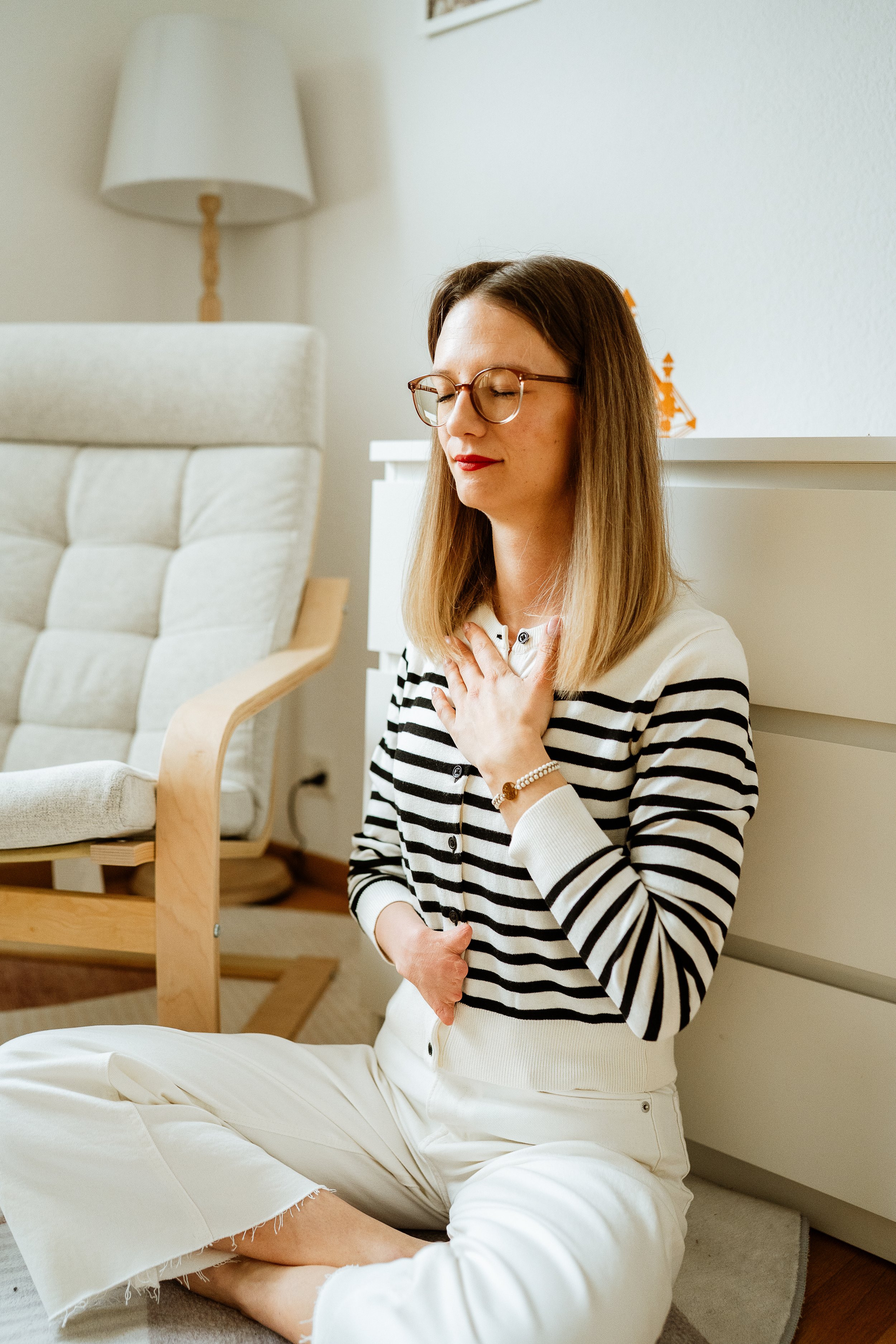 Eine Frau sitzt mit geschlossenen Augen im Schneidersitz auf dem Boden, die rechte Hand auf der Brust und die linke Hand im Schoß, in einem hell eingerichteten Raum.