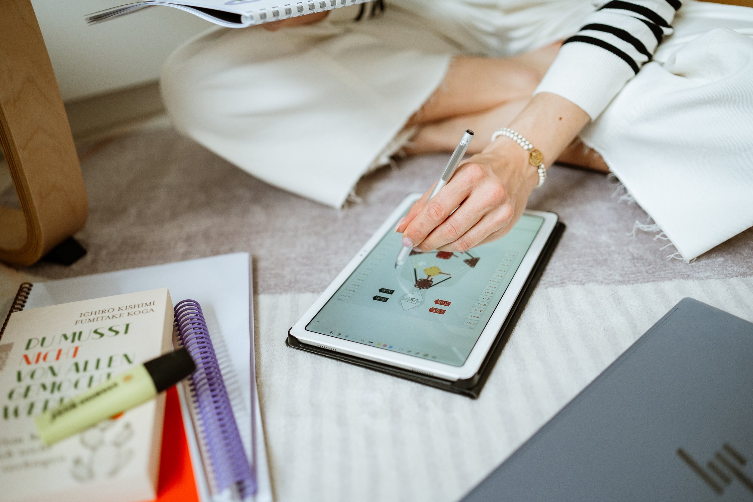 Eine Person arbeitet mit einem digitalen Stift auf einem iPad, um eine Illustration zu zeichnen, umgeben von Büchern, Notizbüchern und Schreibutensilien auf dem Boden.