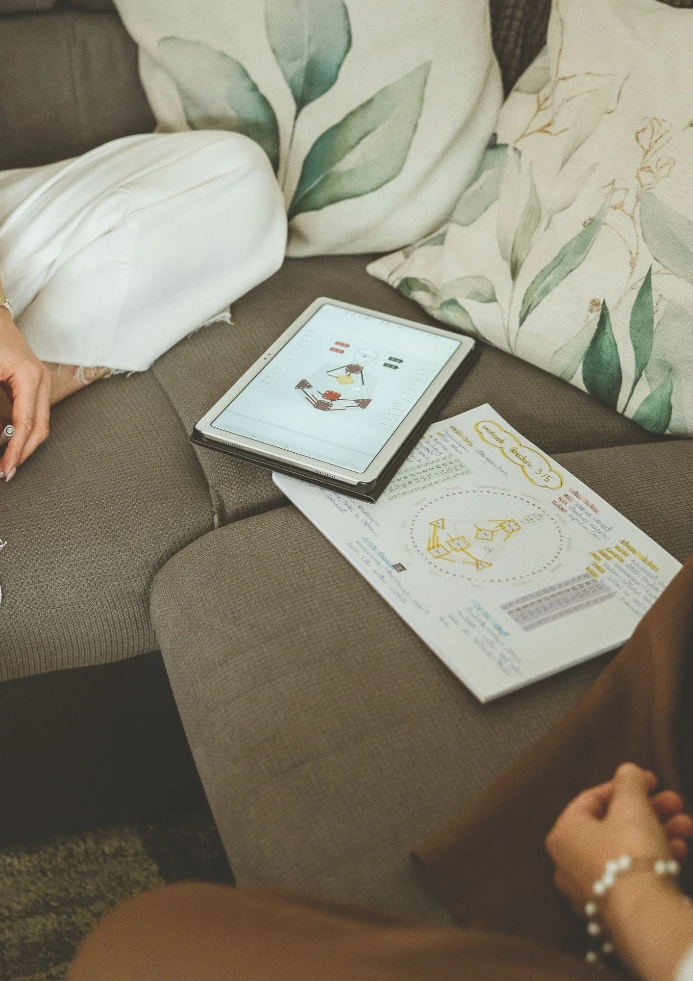 Zwei Personen sitzen auf einem Sofa, während sie an einem Tablet und einem Kursbuch arbeiten, das sich auf den Kurs 'Creative Addiction 3.5' bezieht, mit Zeichnungen und Notizen.