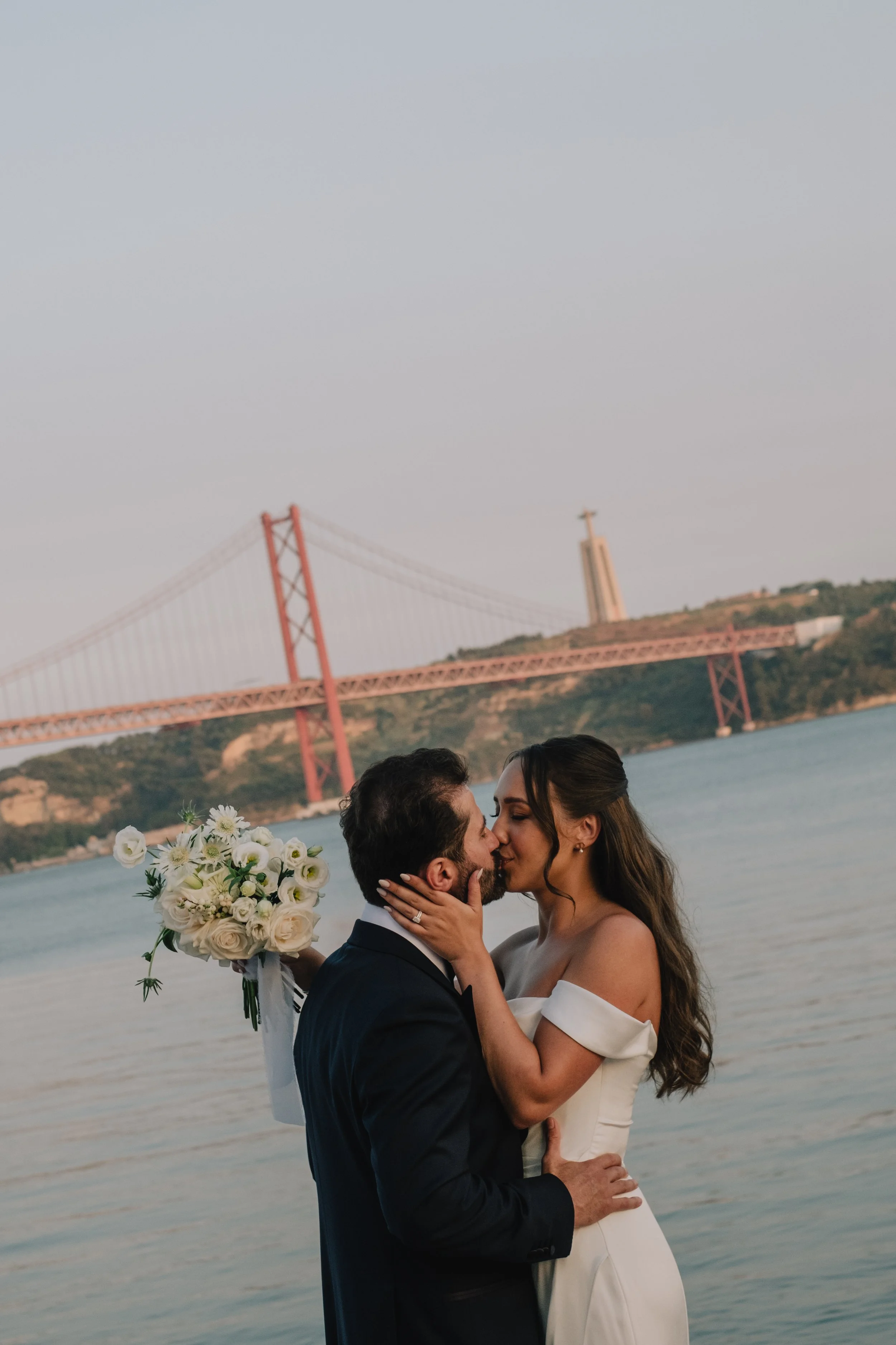Bride and groom kissing by the river with the 25 de Abril bridge in Lisbon during a wedding