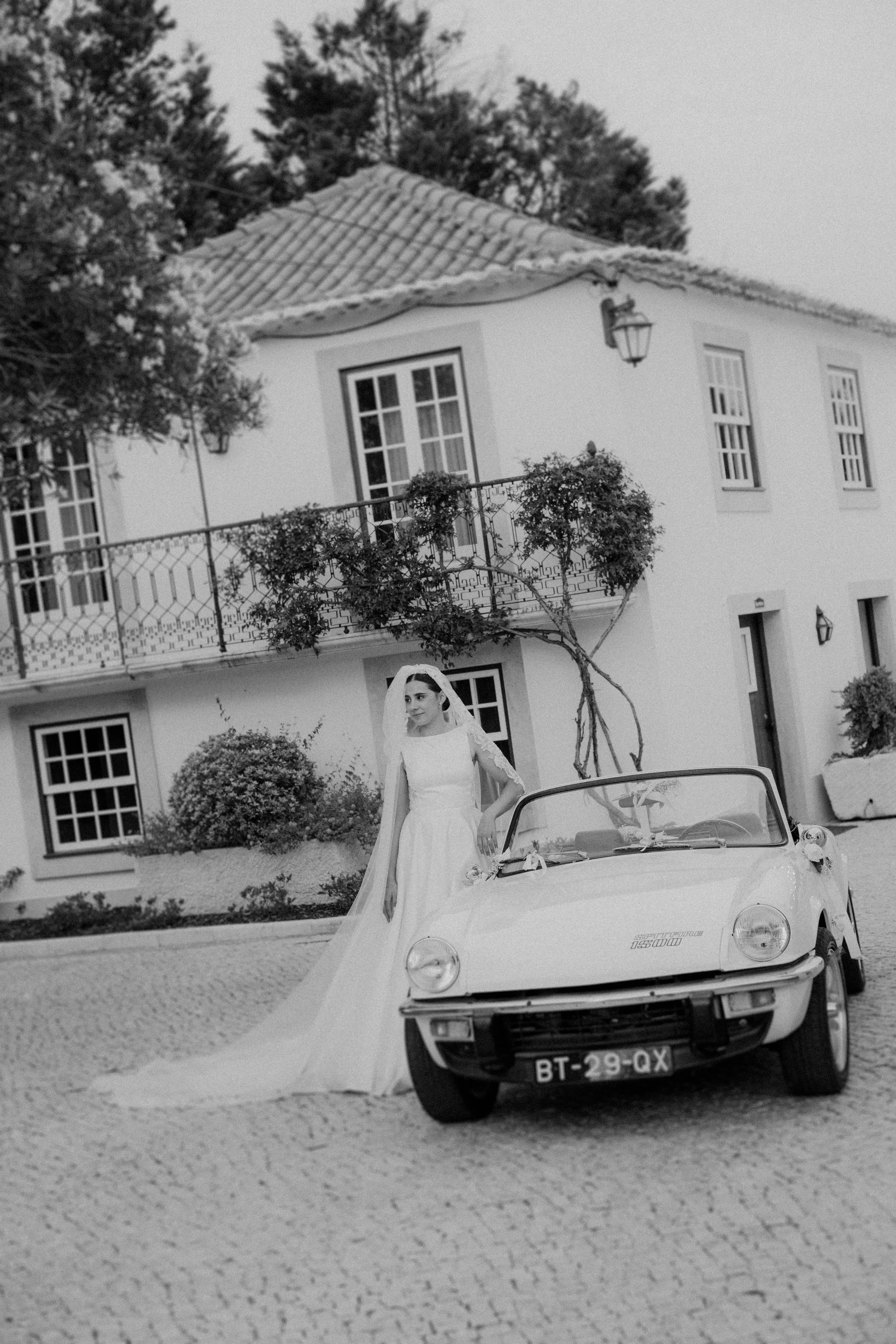 Bride standing next to a vintage car in front of a traditional villa in Portugal captured in black and white