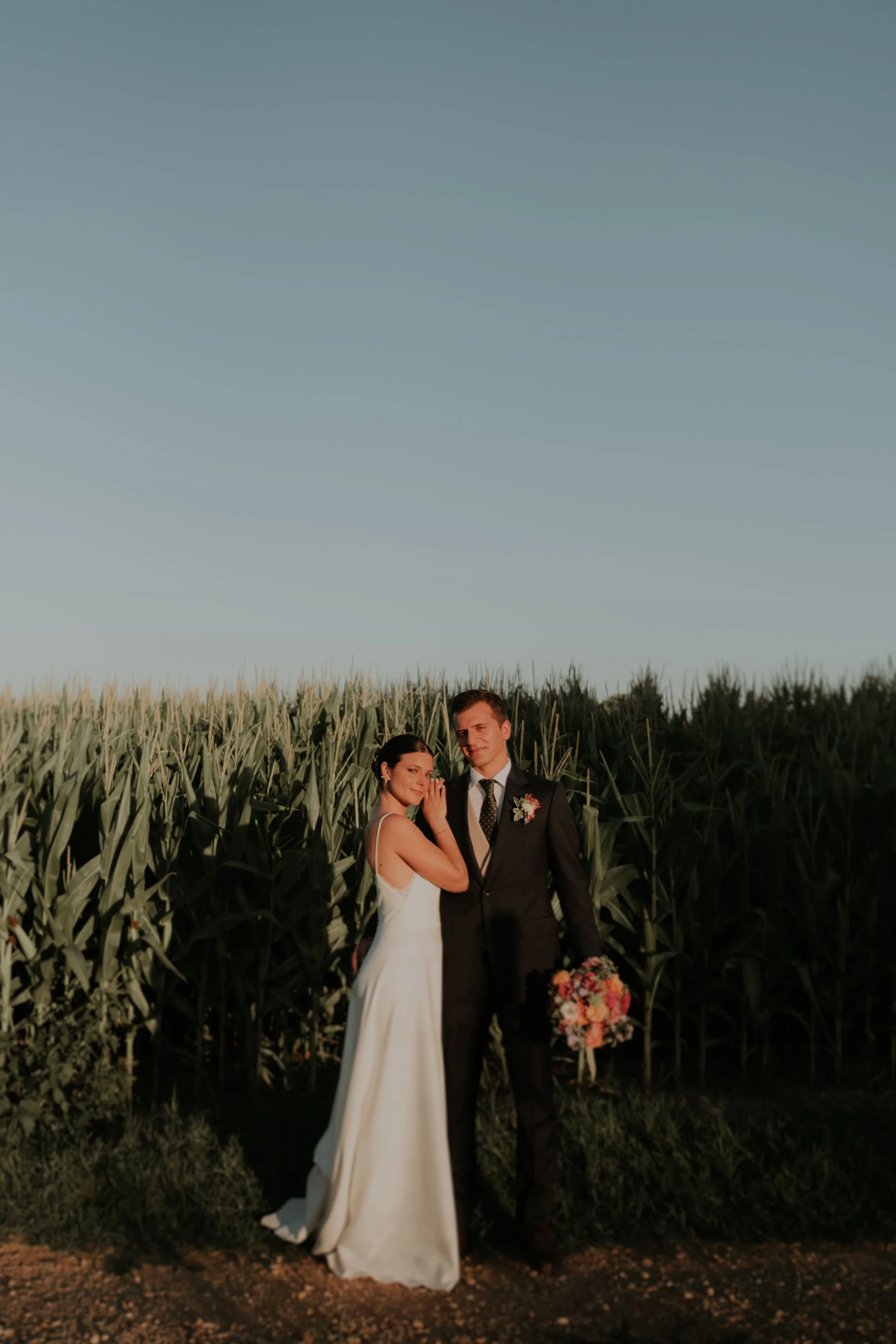 Bride and groom standing side by side in front of a cornfield during golden hour, with soft natural light and clear sky.