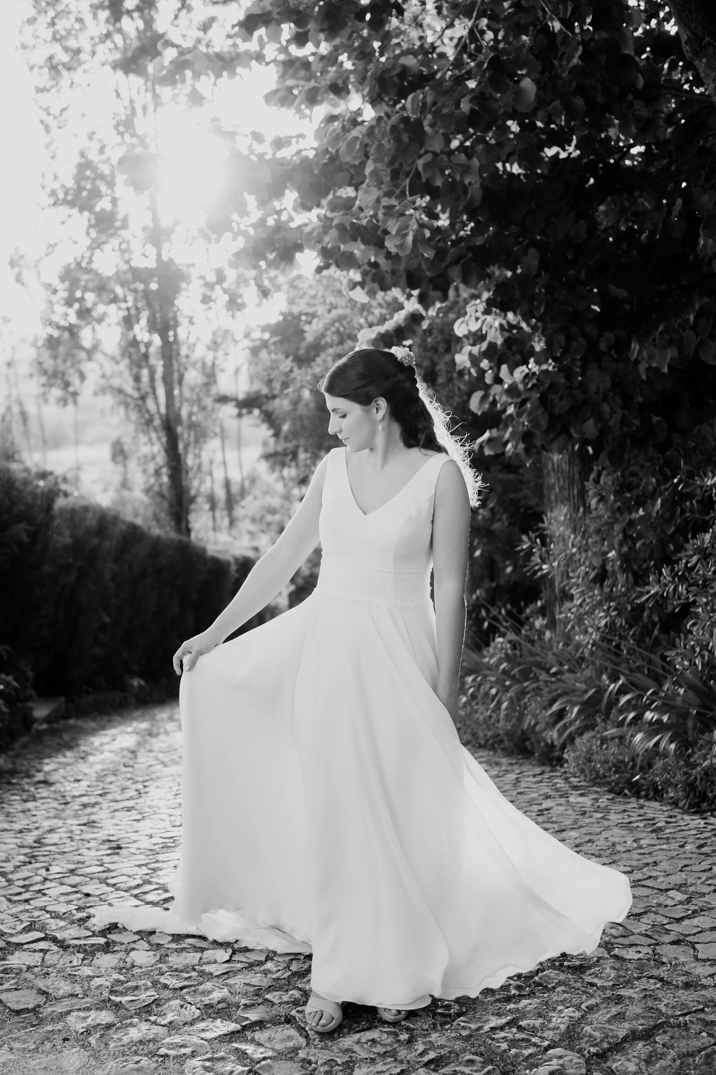 Black and white photo of a bride gently holding her wedding dress while walking on a cobblestone path surrounded by trees, captured during golden hour by Carlos Mendes Photography.