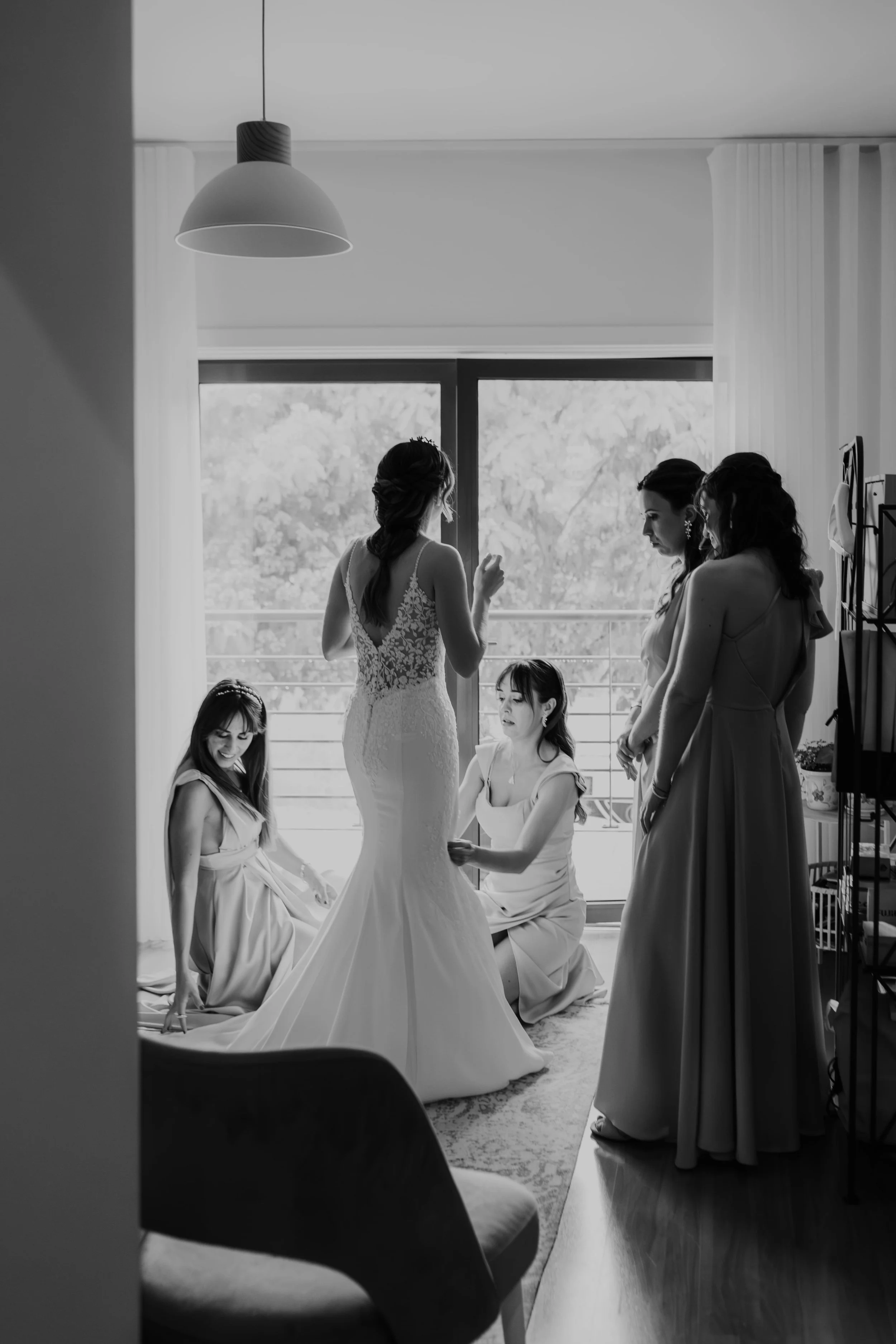 Black and white photograph of a bride surrounded by her bridesmaids while getting ready on the wedding day, captured in a softly lit room with a large window, by Carlos Mendes Photography.
