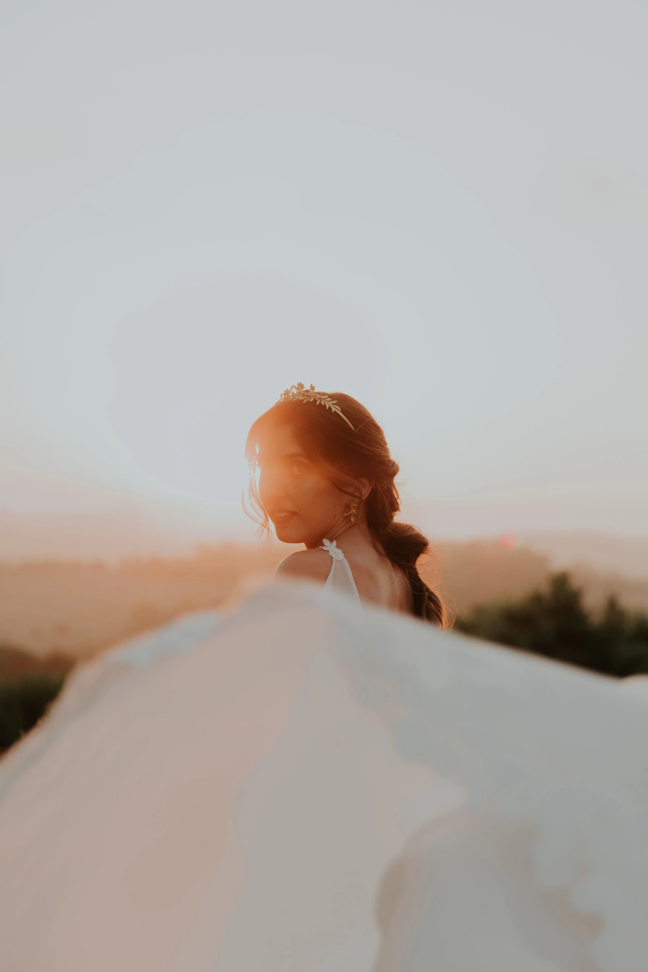 Close-up portrait of a bride bathed in sunset light, wearing a delicate tiara and soft veil in the foreground, with a dreamy golden-hour atmosphere, captured by Carlos Mendes Photography.
