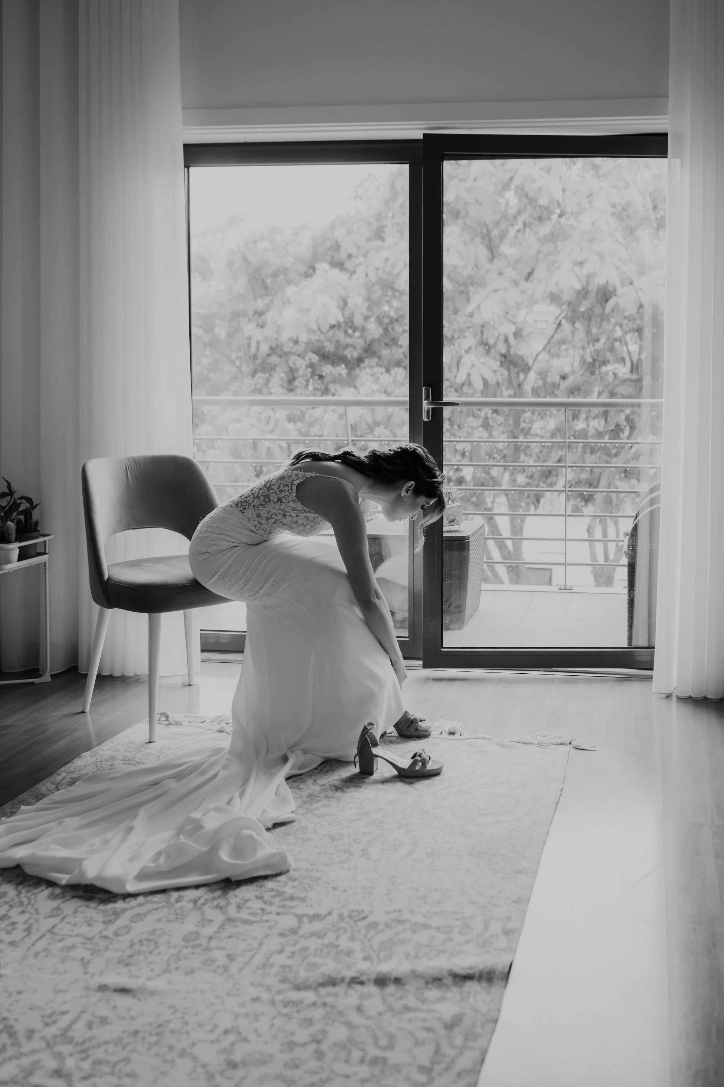 Black and white photo of a bride sitting on a chair and putting on her wedding heels, captured in soft natural light near a large window, by Carlos Mendes Photography.