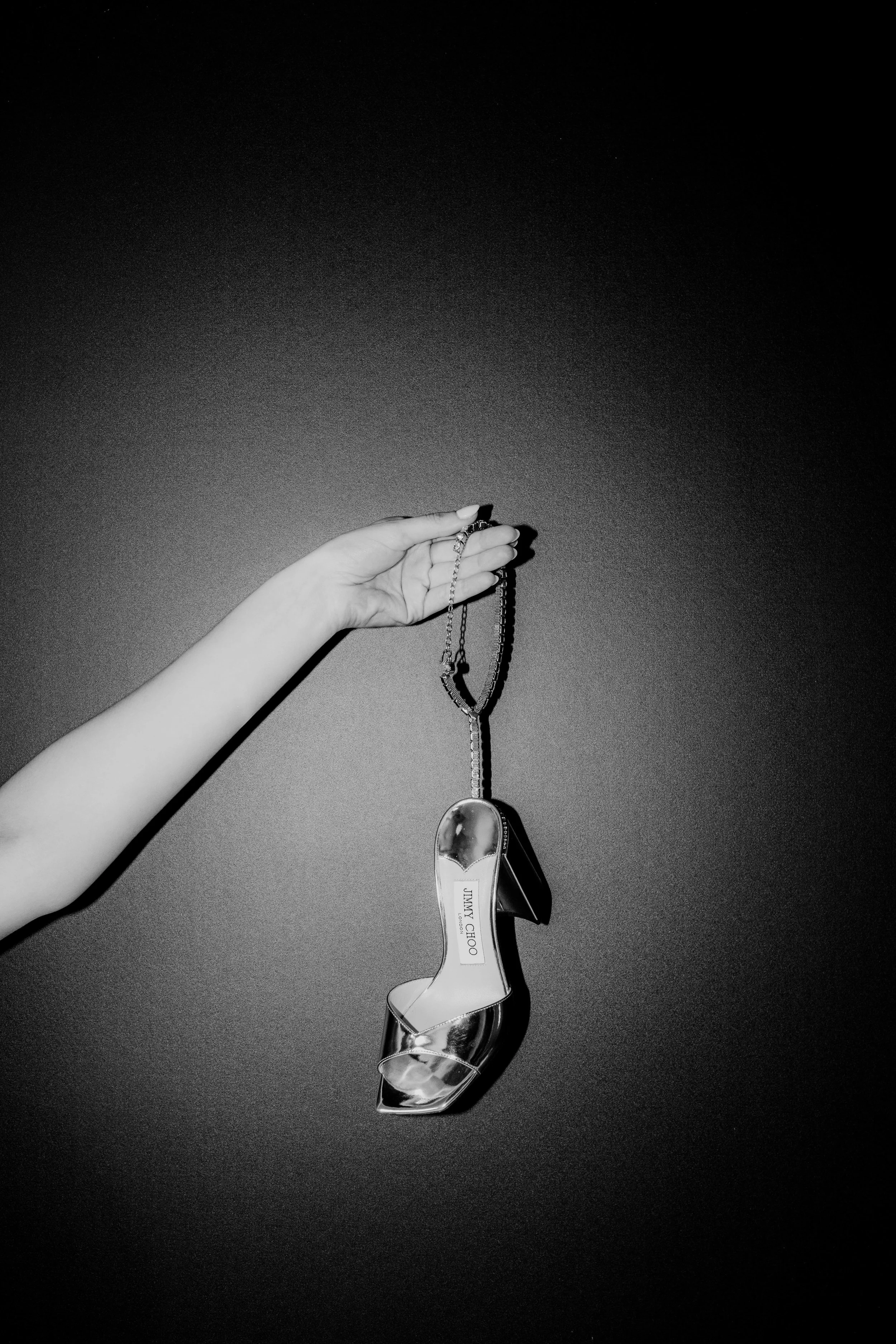 Black and white photograph of a female hand holding a metallic Jimmy Choo high heel by a delicate chain against a dark textured background, captured by Carlos Mendes Photography.