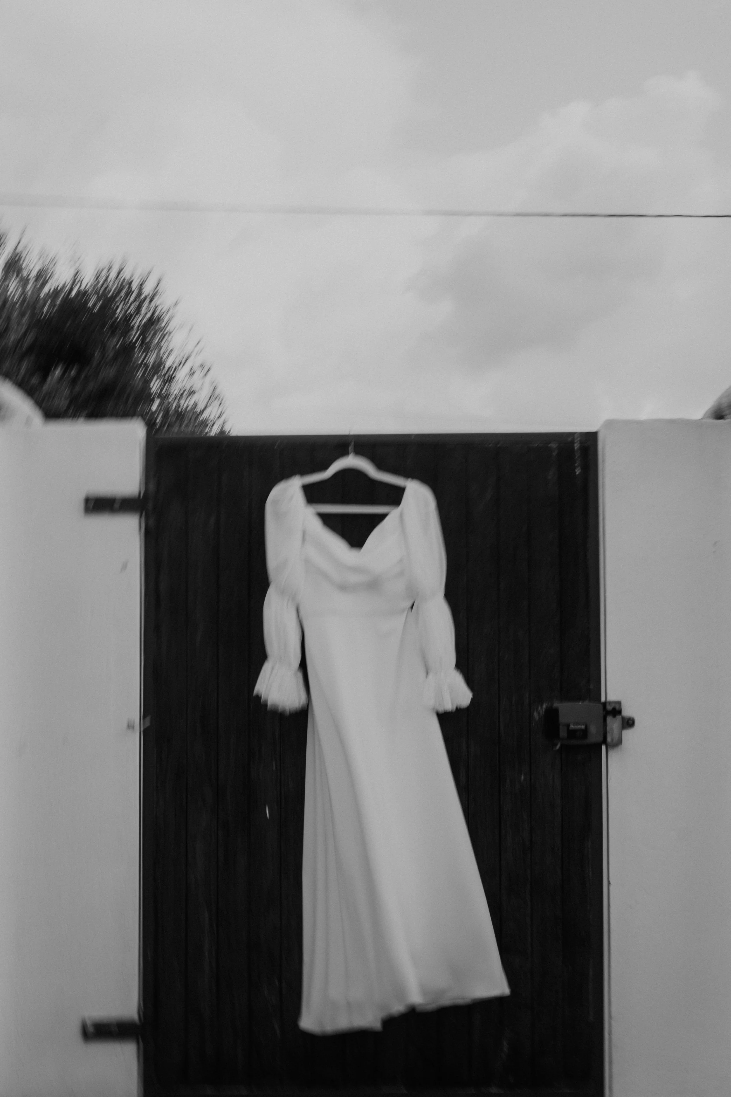 Black and white photo of a wedding dress with puff sleeves hanging on a wooden gate, captured outdoors.
