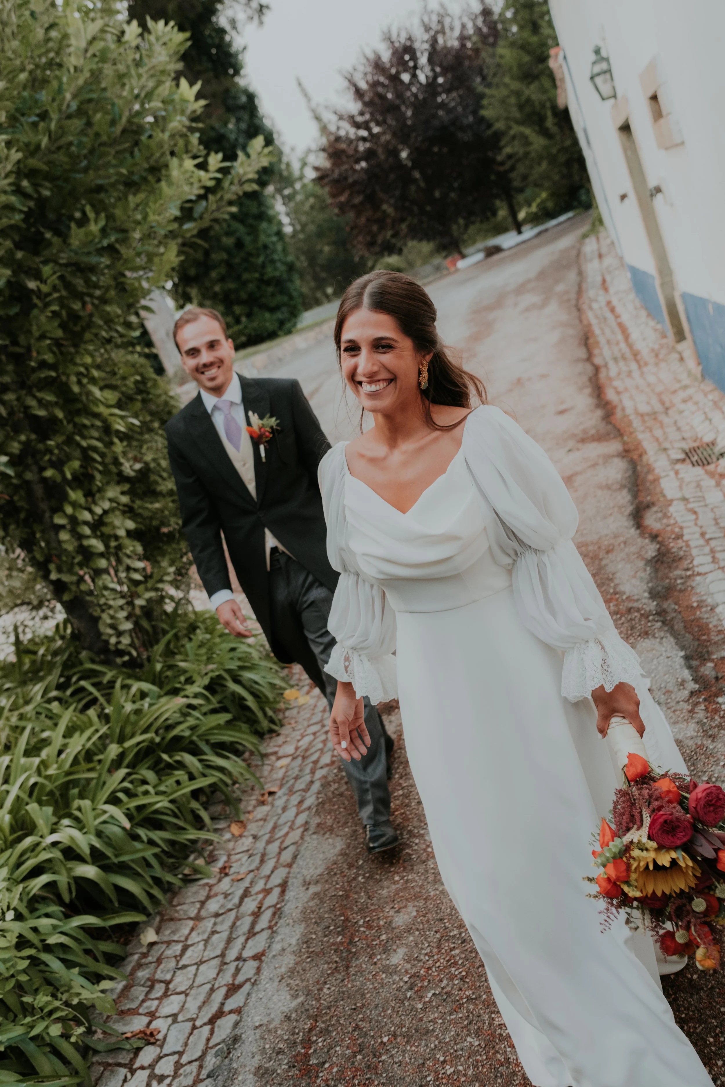 Bride and groom walking joyfully along a garden path, with the bride smiling brightly and holding a colorful bouquet.