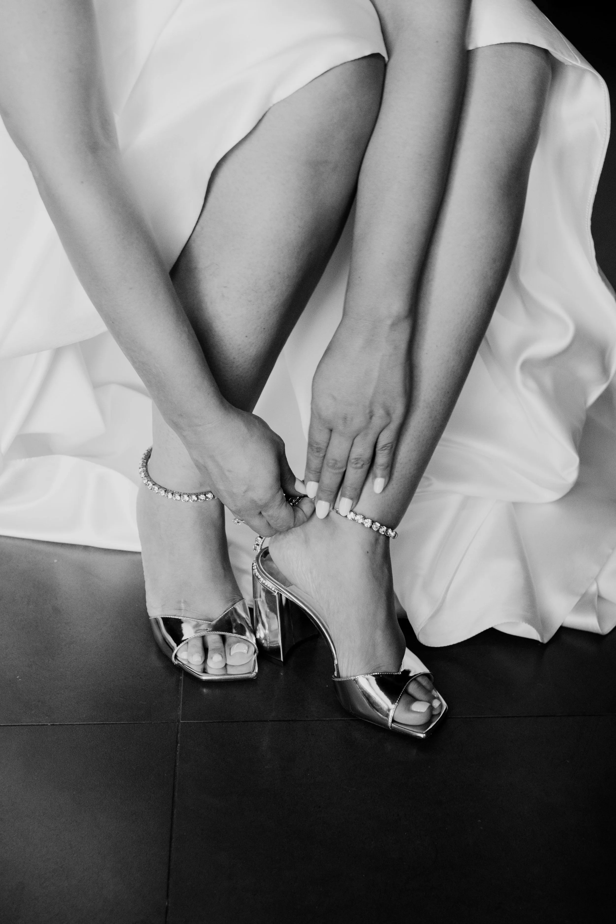 Black and white photo of a bride fastening a diamond anklet and wearing metallic block heels, showcasing bridal elegance and wedding morning details, captured by Carlos Mendes Photography.