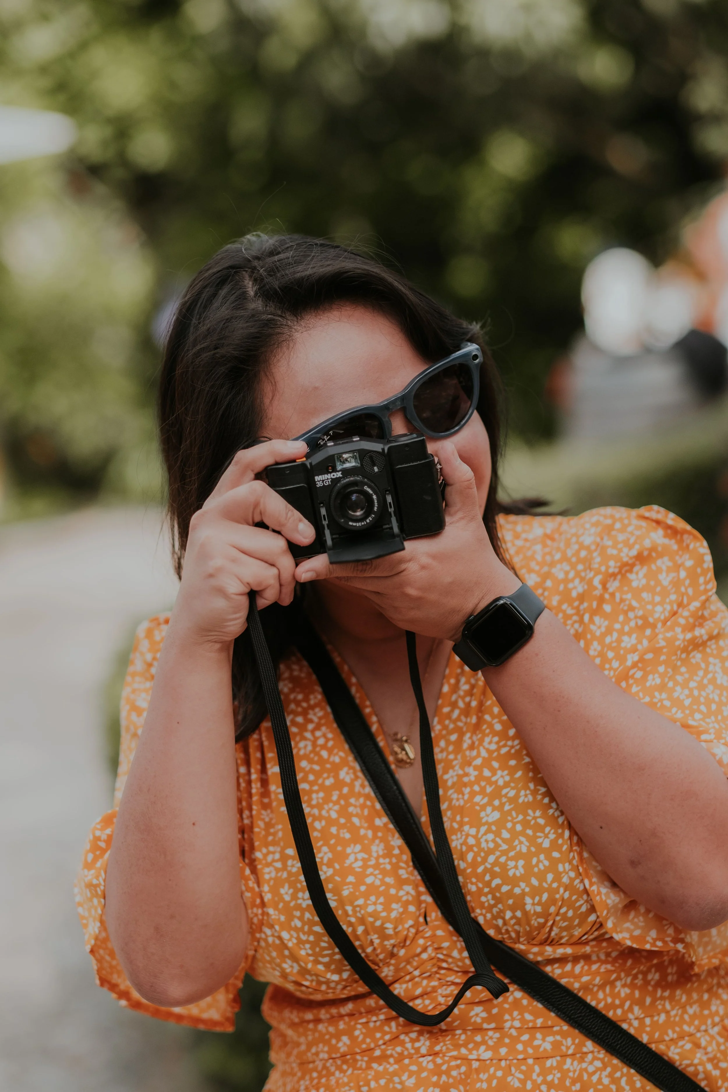 Wedding guest in a yellow floral dress taking a photo with a vintage film camera during an outdoor celebration. Captured by Carlos Mendes Photography.