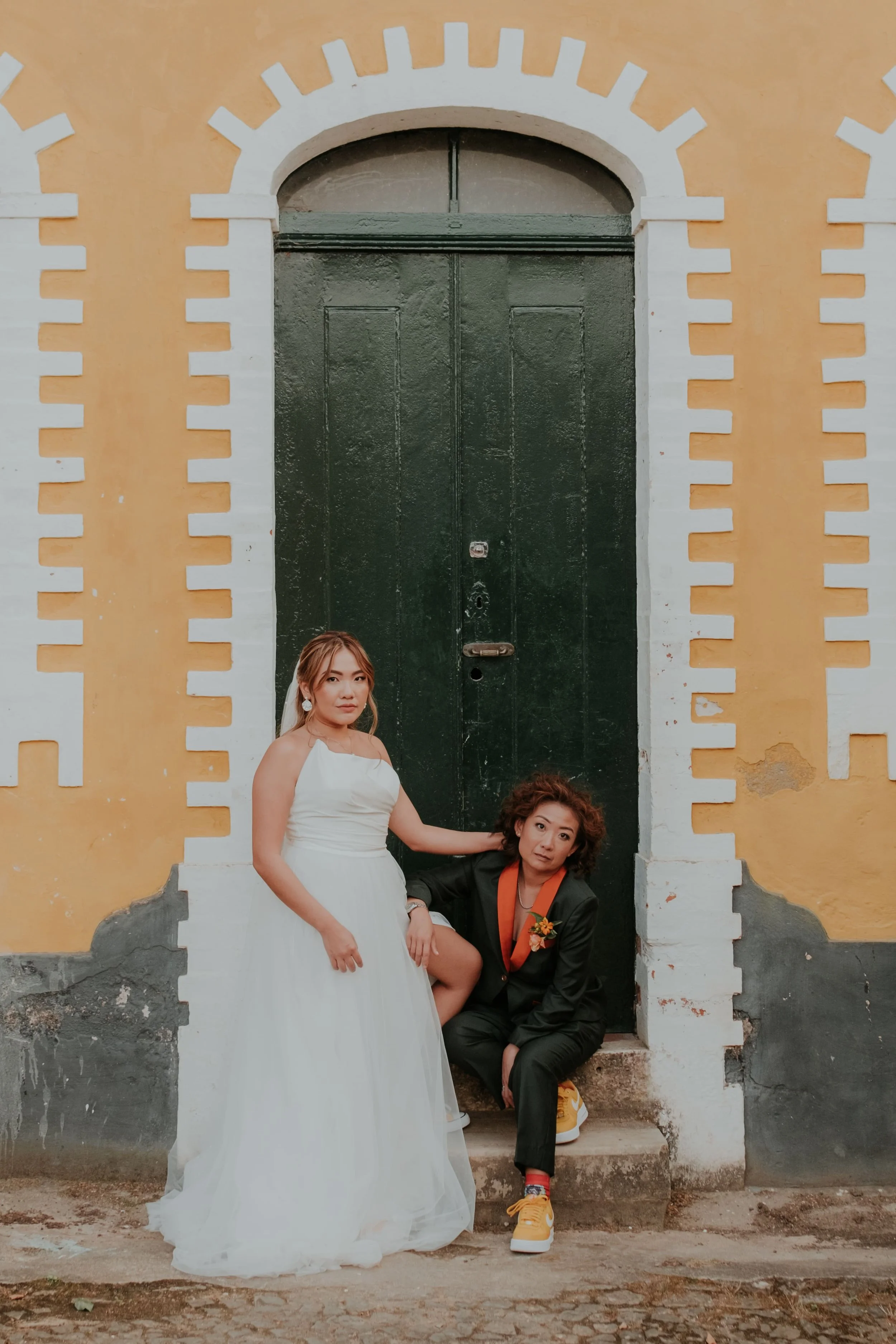 Stylish brides posing in front of a colorful yellow and green building; one in a wedding dress and the other in a suit with orange sneakers, captured by Carlos Mendes Photography.