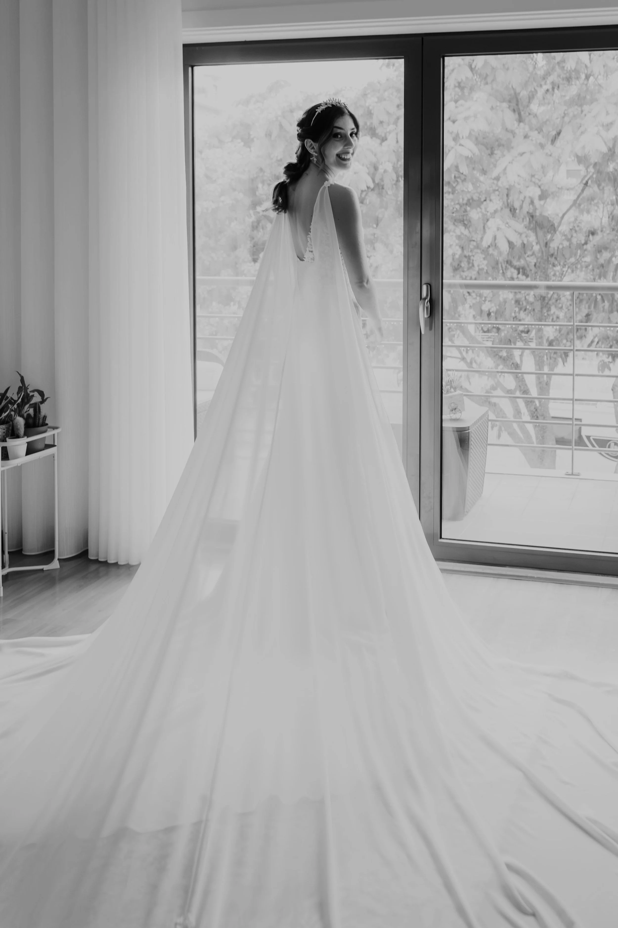 Black and white photo of a smiling bride in a long train wedding gown standing by a large window, illuminated by soft natural light, captured by Carlos Mendes Photography.