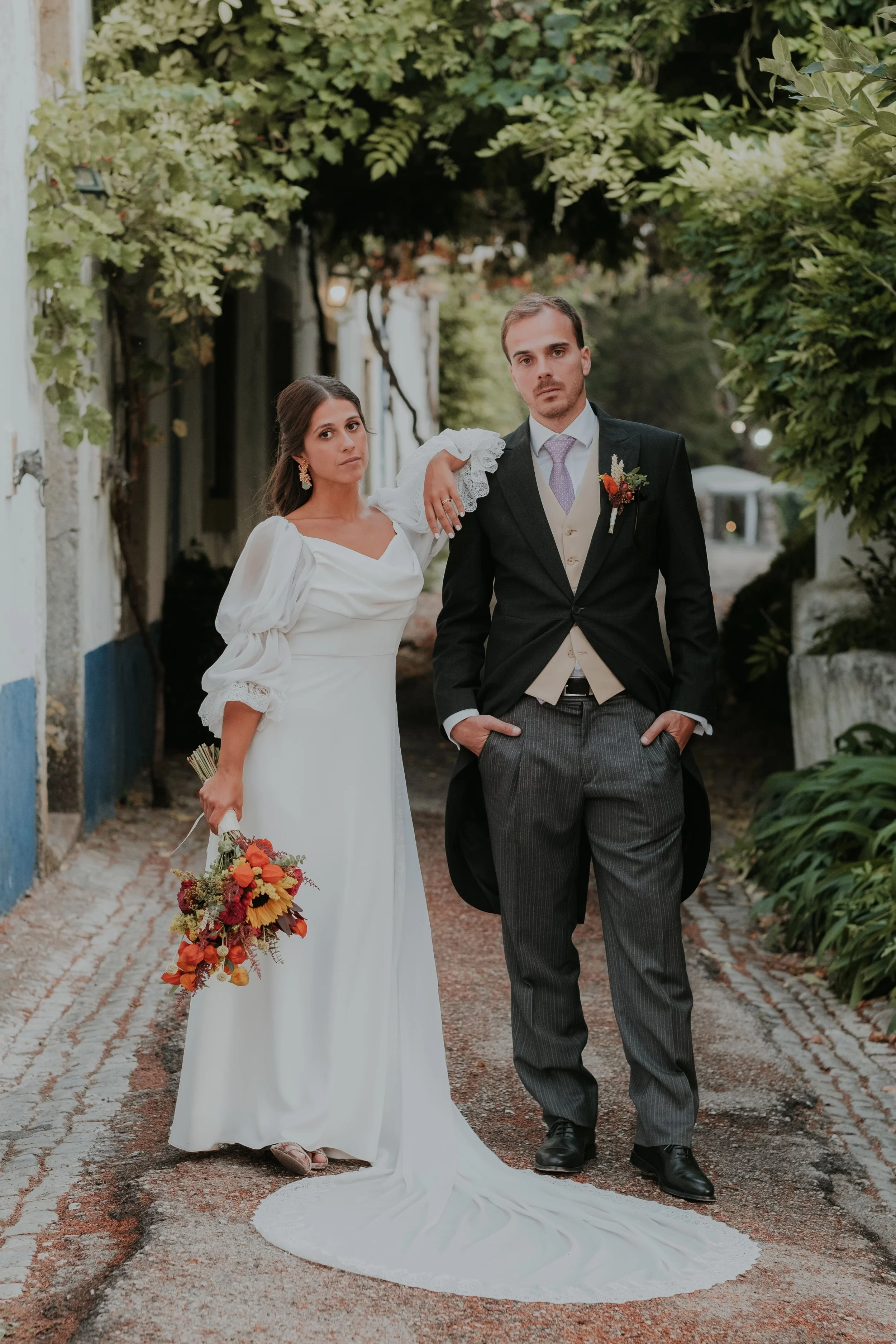 Bride and groom posing elegantly on a rustic garden path, with the bride holding a vibrant bouquet and resting her arm on the groom's shoulder.